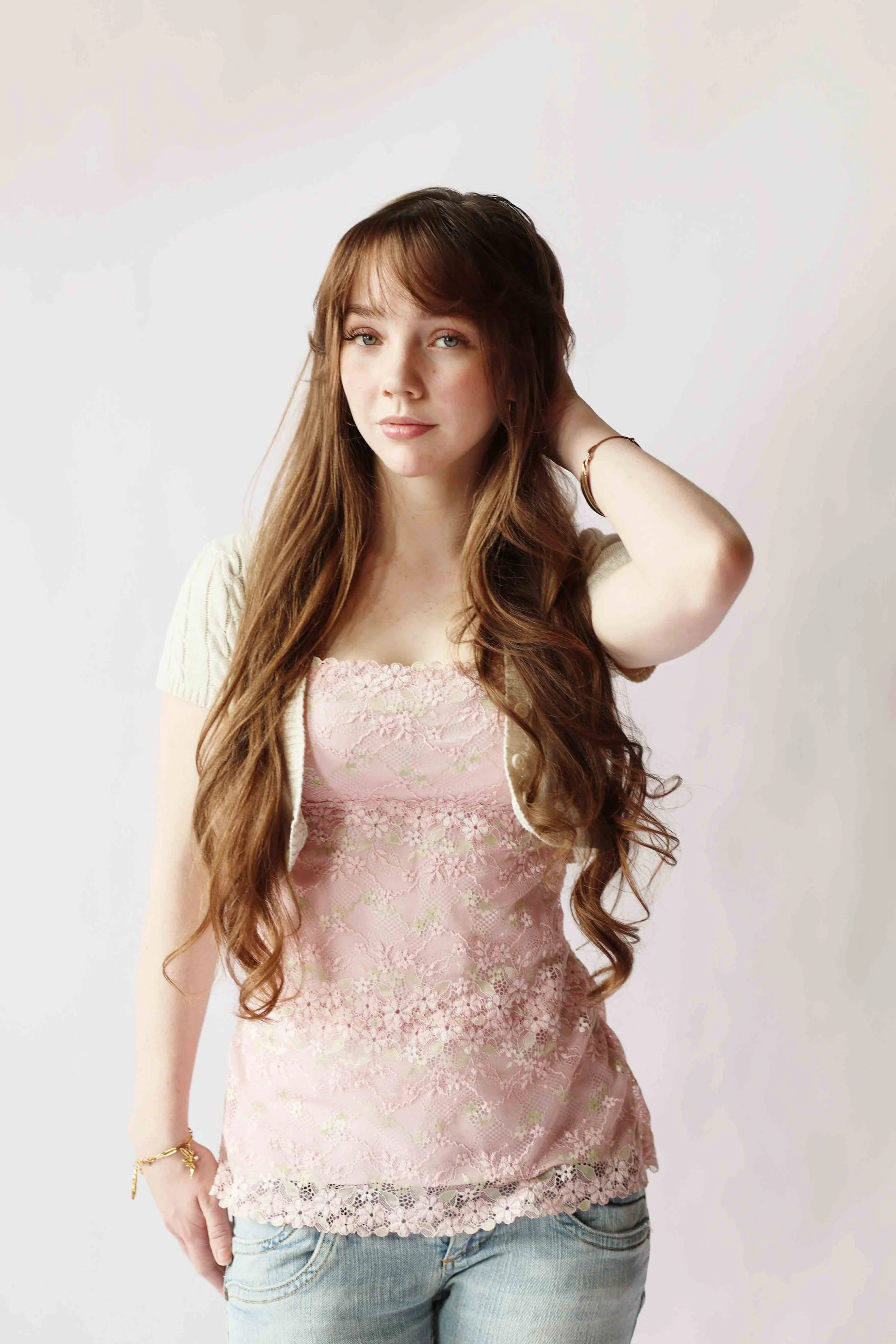 A young woman with long wavy brown hair, light skin, and green eyes, touching her hair, wearing a light pink lace top, denim jeans, and gold jewelry, standing against a plain white background.