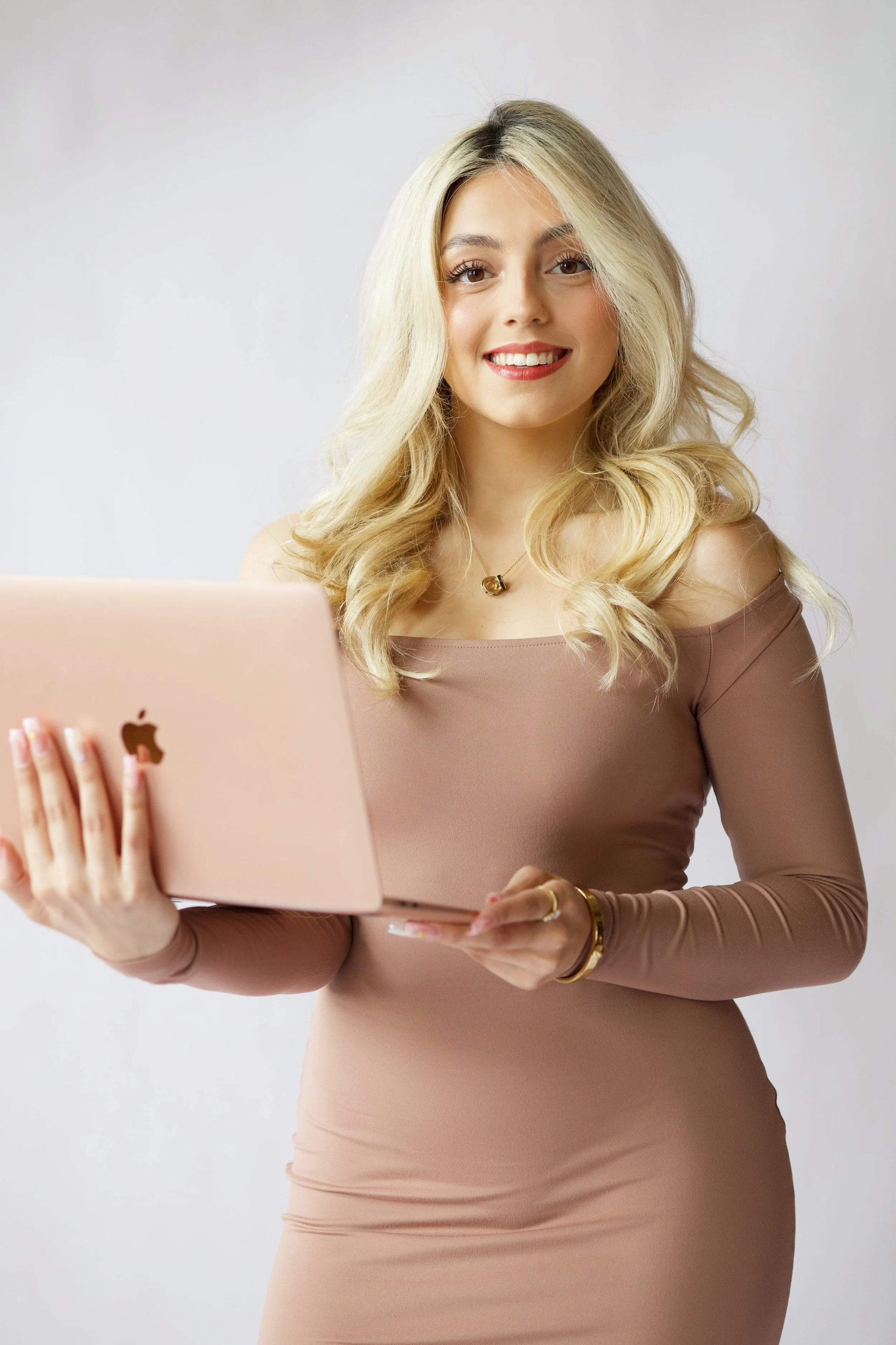 A woman with long blonde hair wearing a beige long-sleeve dress, holding a pink laptop and smiling at the camera. She is standing against a plain light-colored background.