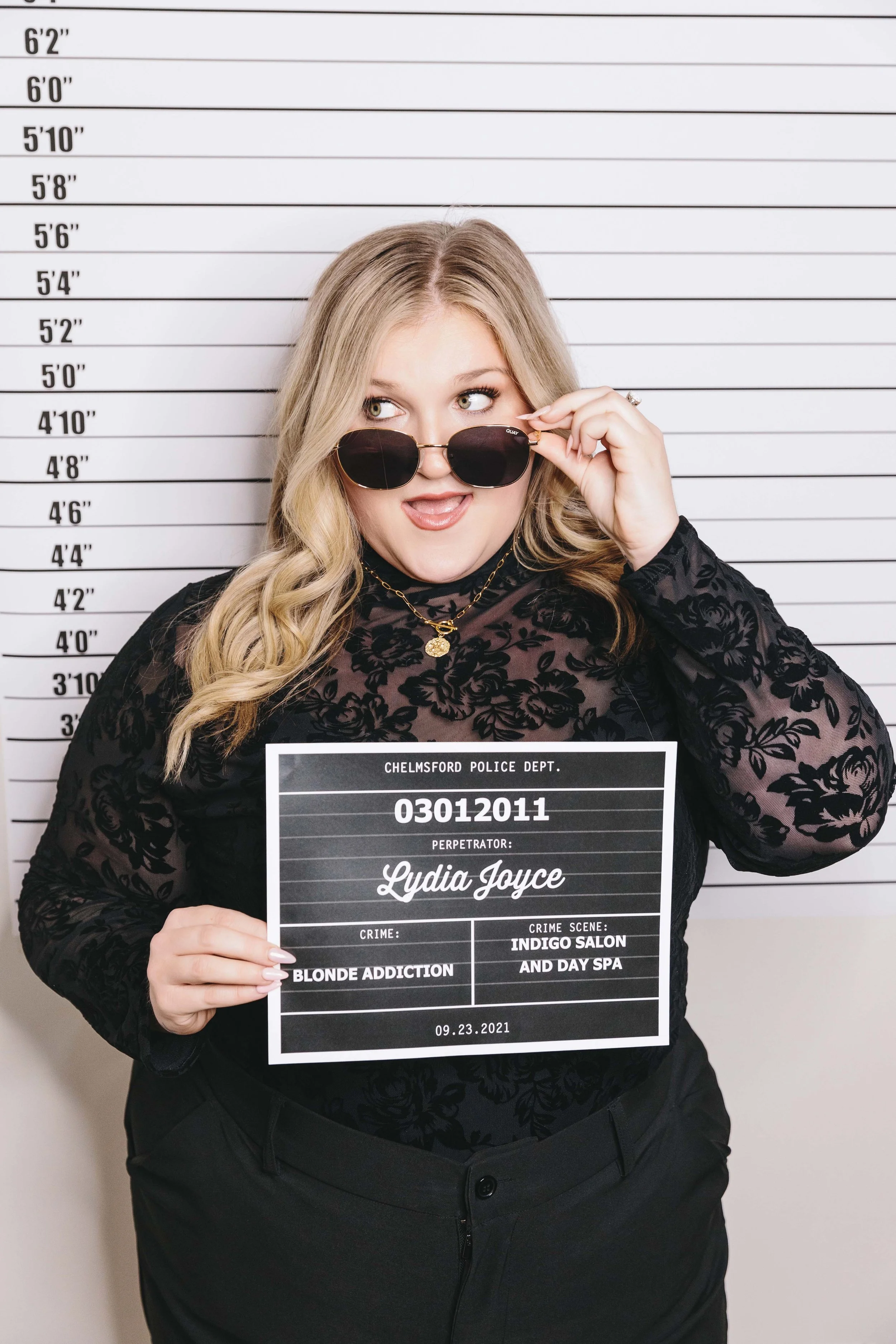 A woman with long blonde hair wearing sunglasses and a black floral top holding a police mugshot board with her information, standing in front of a height chart.