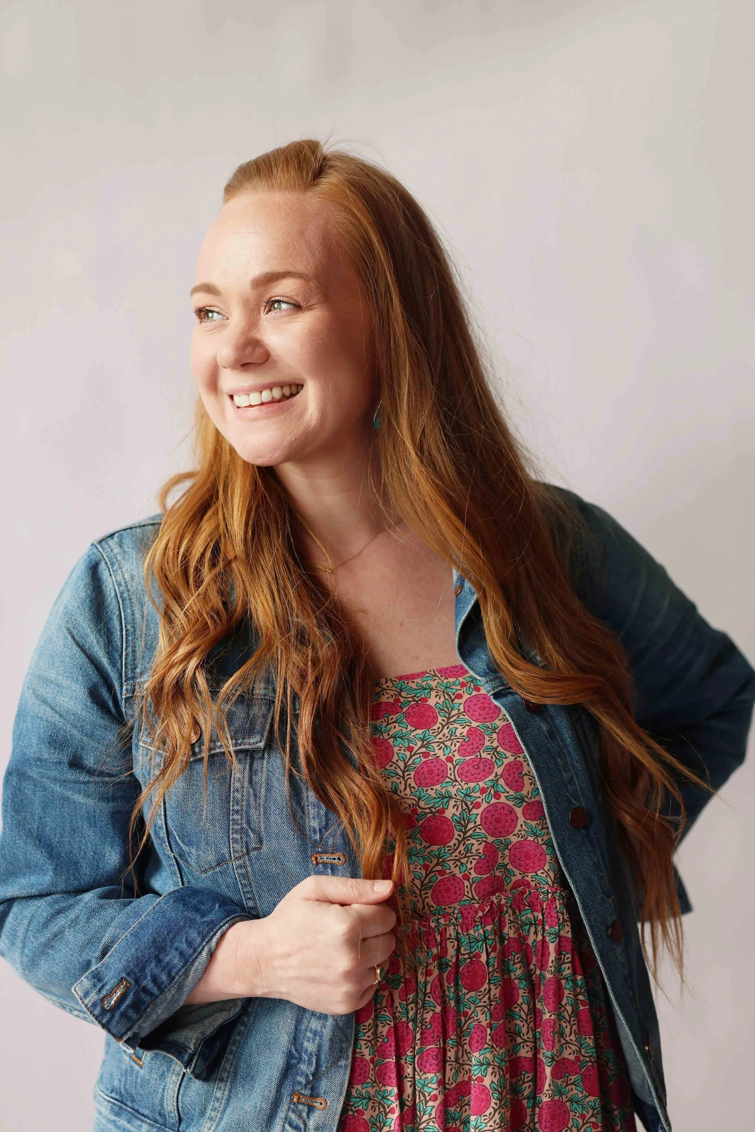 A woman with long red hair, wearing a denim jacket over a colorful dress with berry and leaf patterns, smiling and looking to the side.