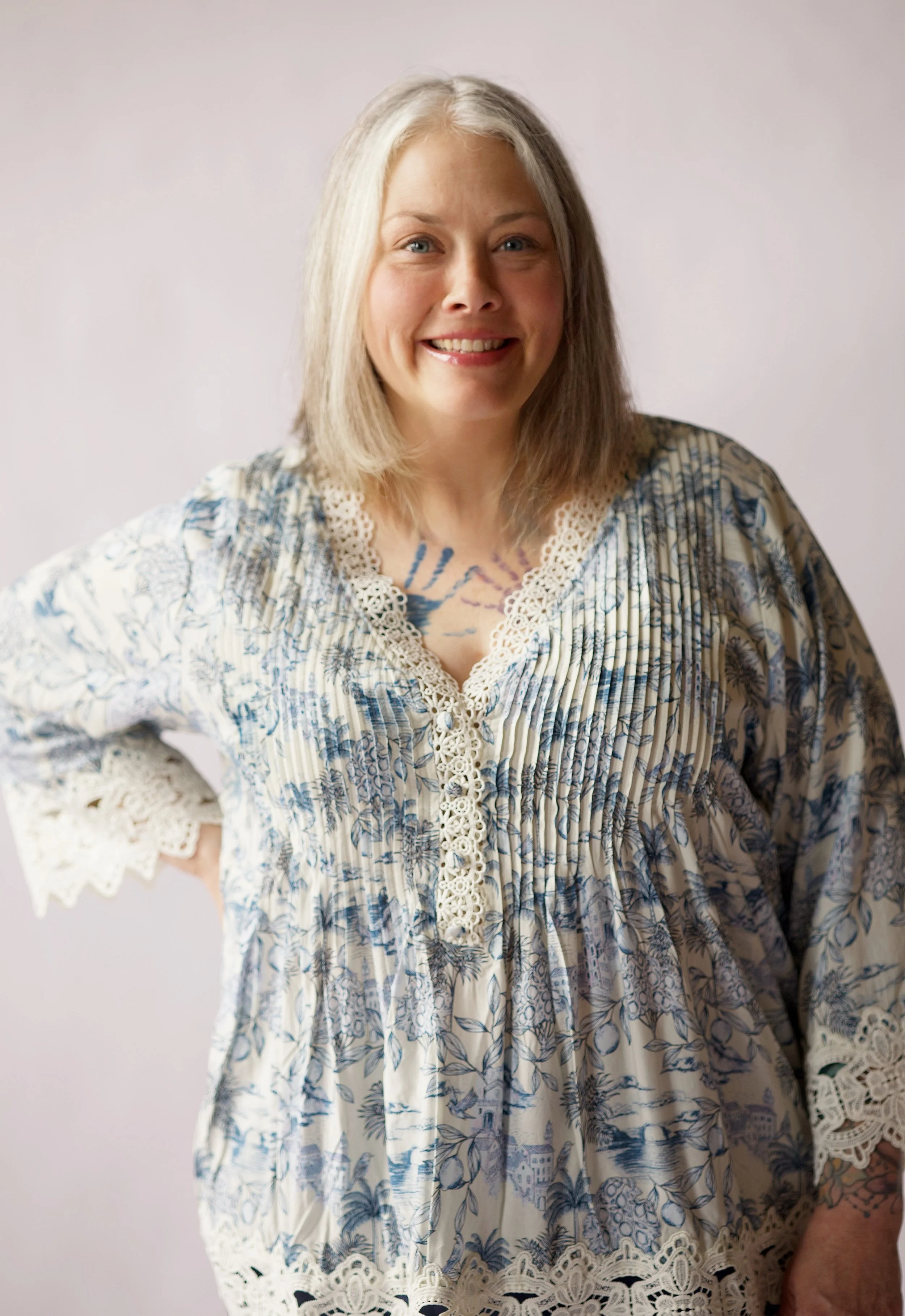 A woman with shoulder-length gray hair, blue eyes, and a wide smile, wearing a cream-colored bohemian-style dress with blue floral patterns and lace accents, standing against a plain light-colored background.