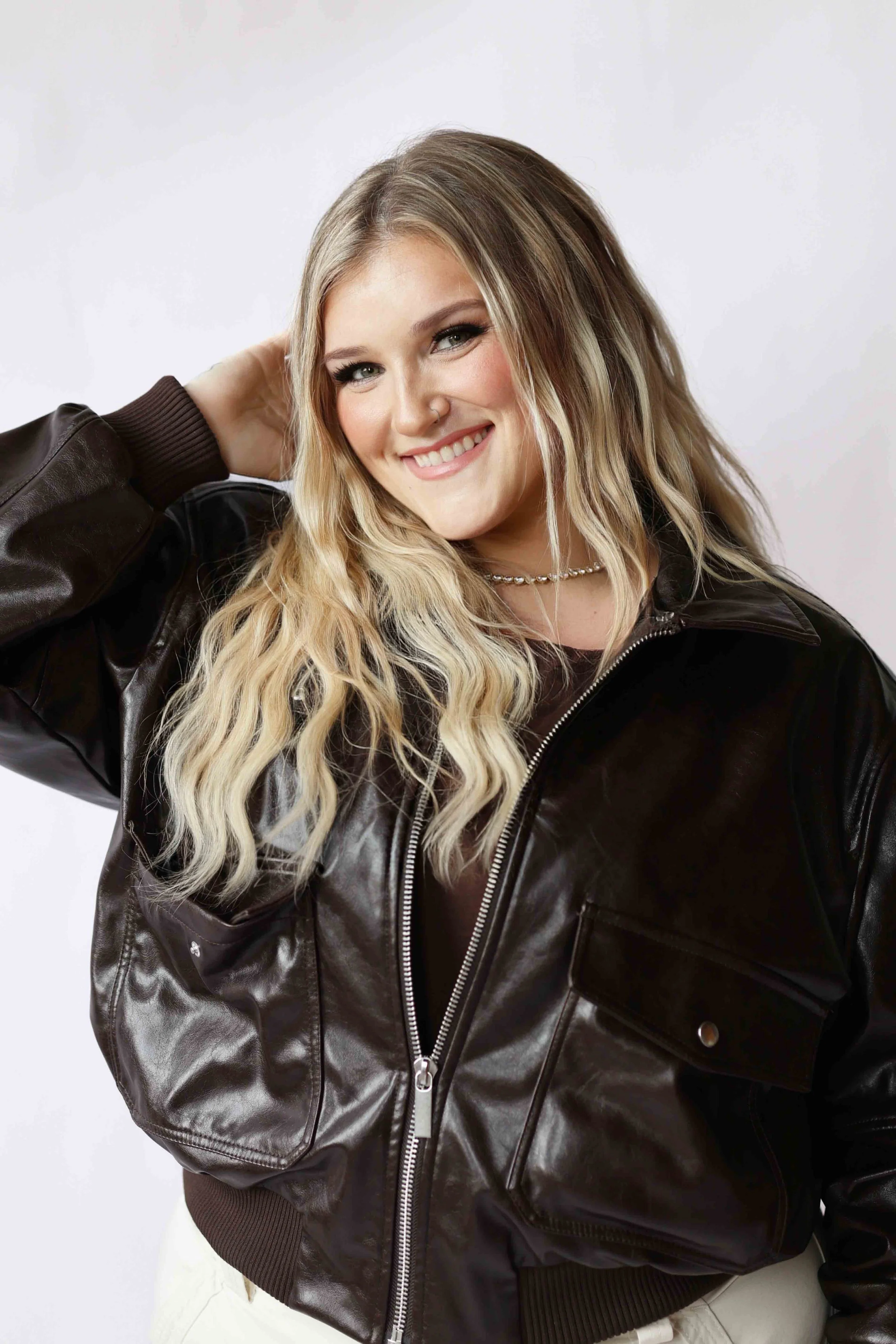 Portrait of a young woman with long blonde wavy hair, wearing a shiny black leather jacket and a pearl necklace, smiling and touching her hair on the back of her head.