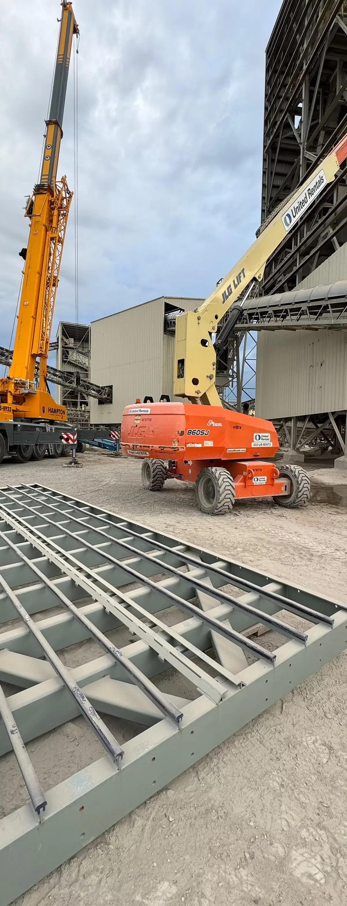 Construction site with orange and yellow telescopic boom lift and crane, metal framework on ground, and industrial buildings in background.