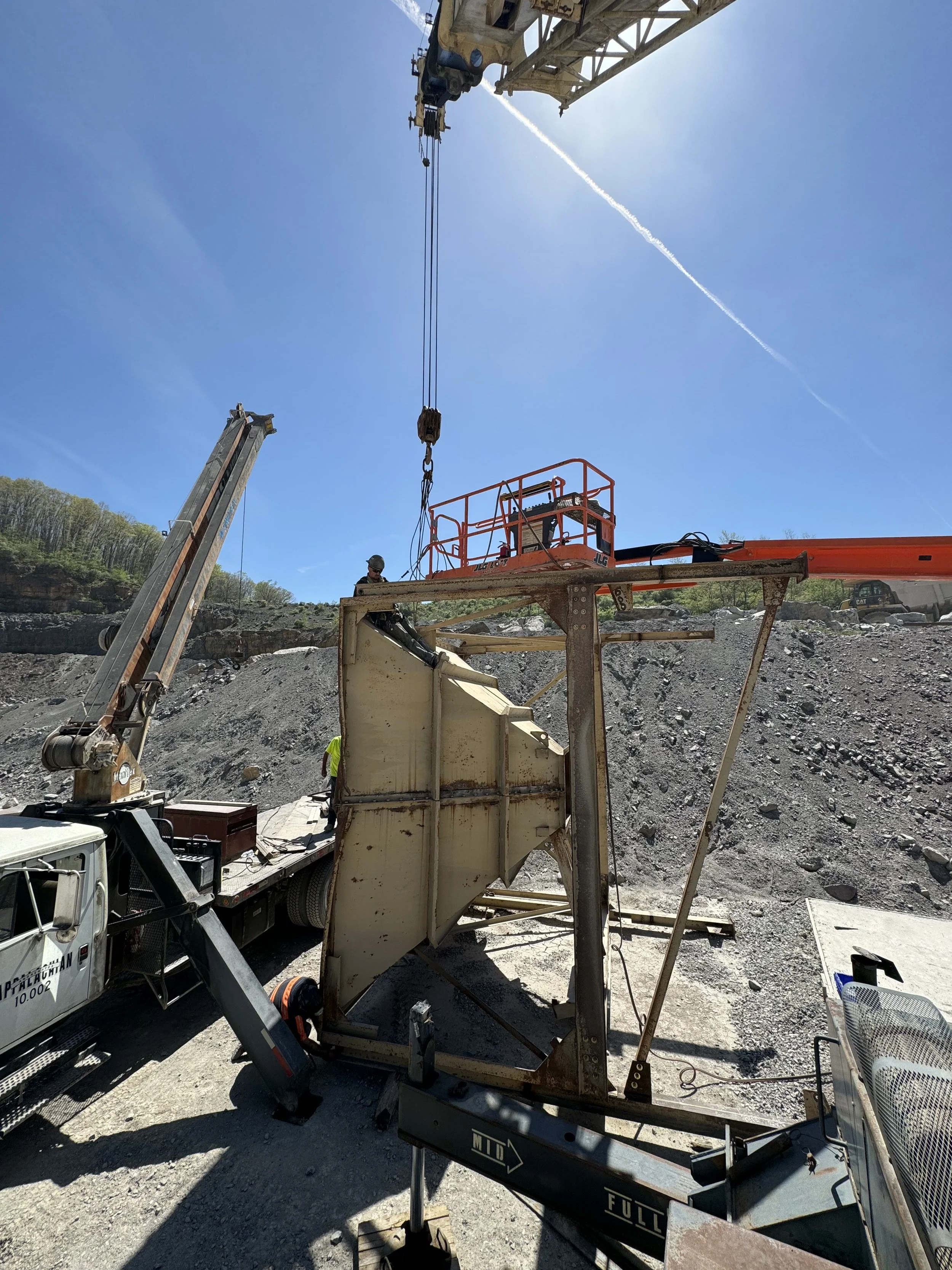 Construction site with a crane lifting a metal structure, workers wearing safety gear, and a rocky landscape under a clear blue sky.