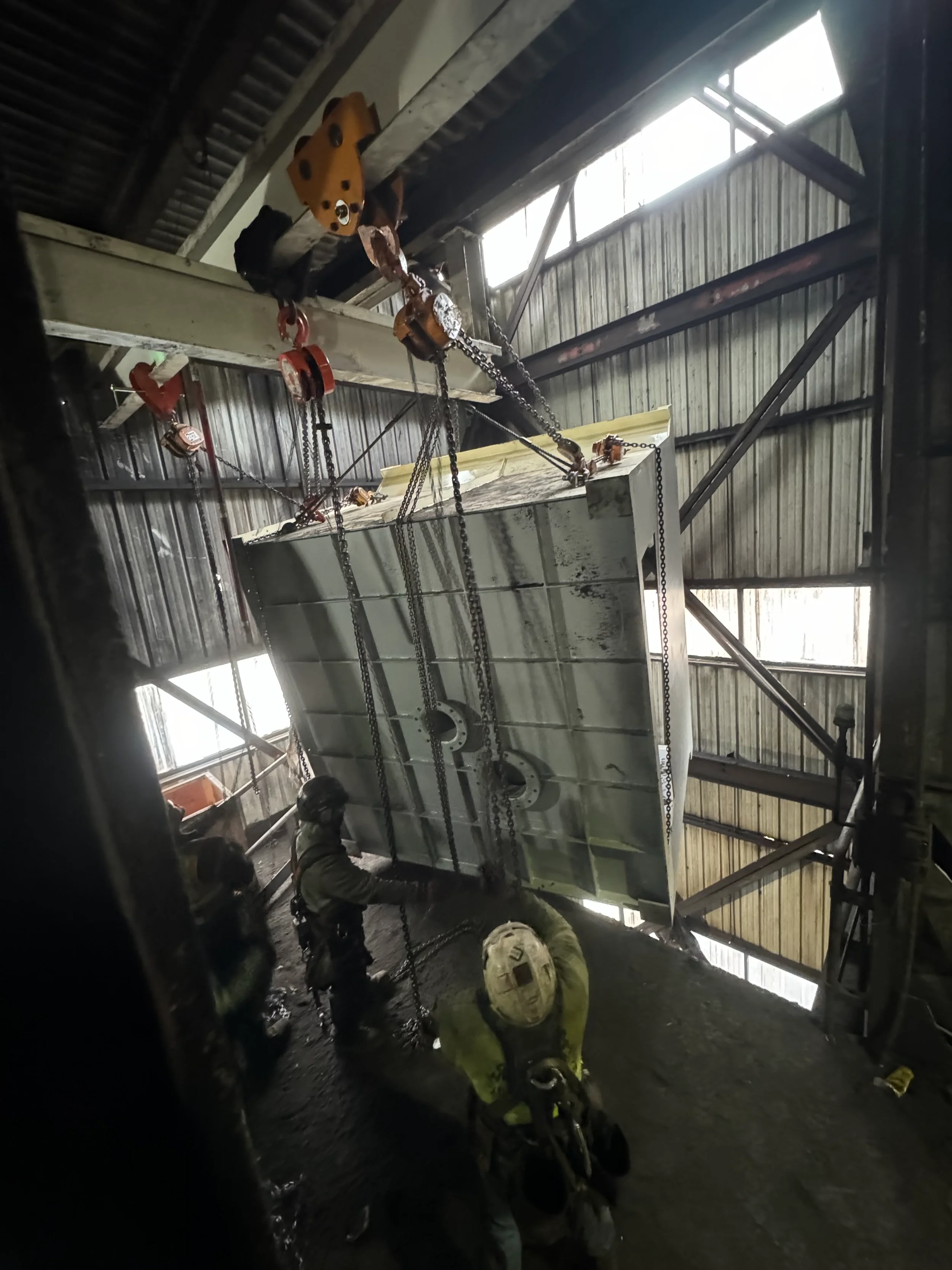 Workers in safety gear lifting a large metal structure with chains inside an industrial building.