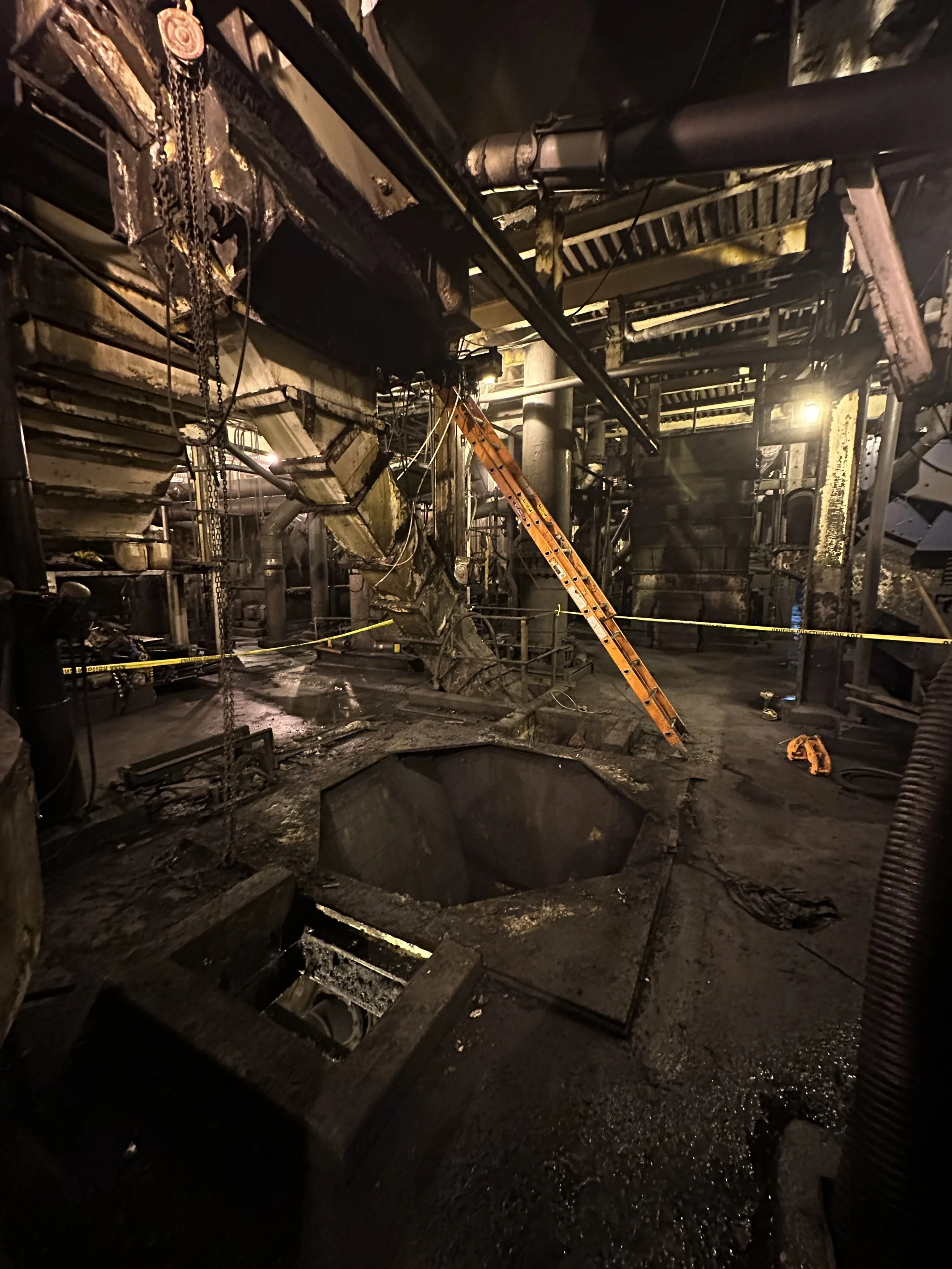 Interior of a building with damaged and charred structures, exposed pipes, and broken flooring, indicating fire damage, with a ladder leaning against the wall and emergency caution tape visible.