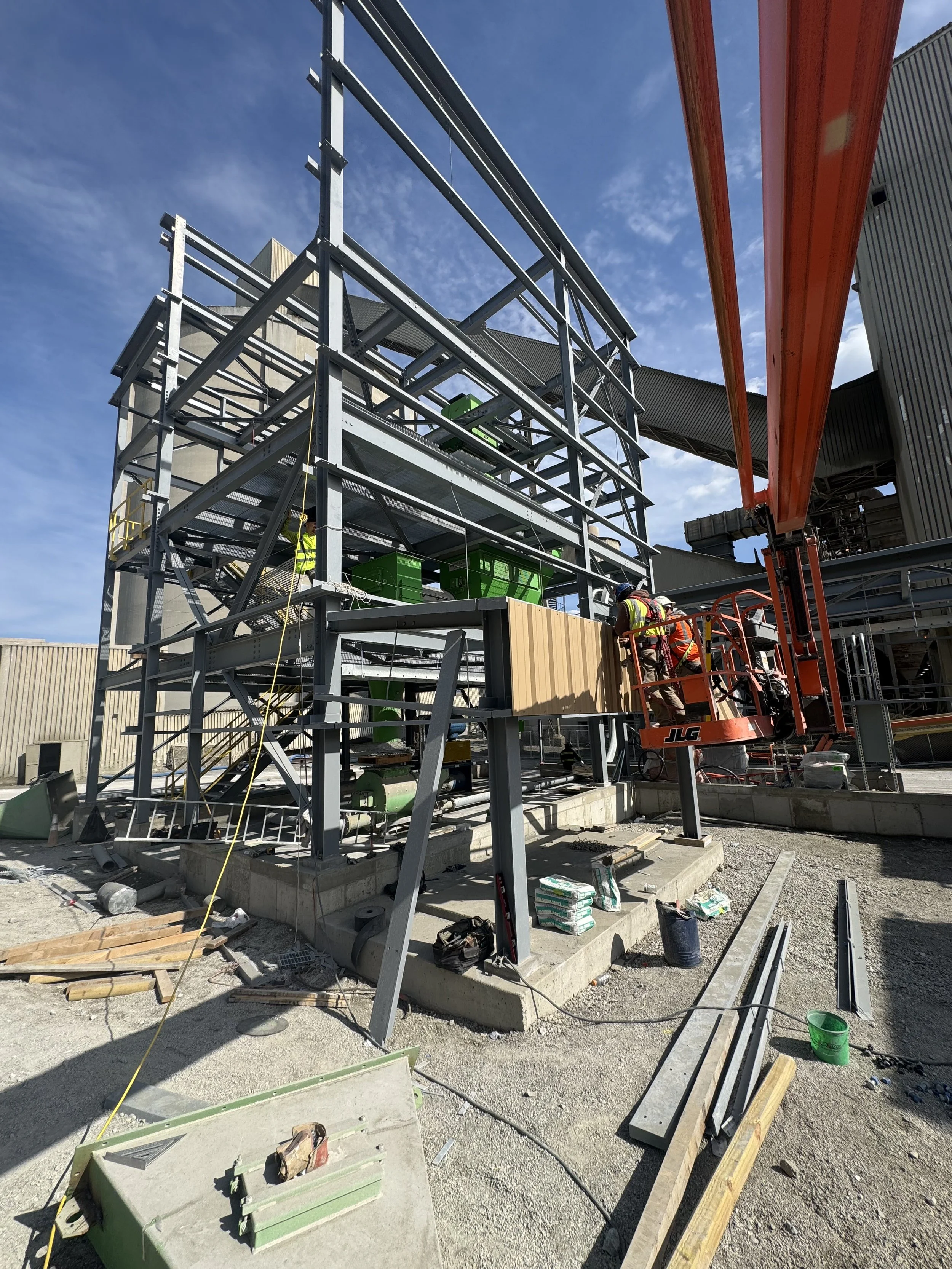 Construction site with steel framework, workers in safety gear, and building materials outdoors under a blue sky.
