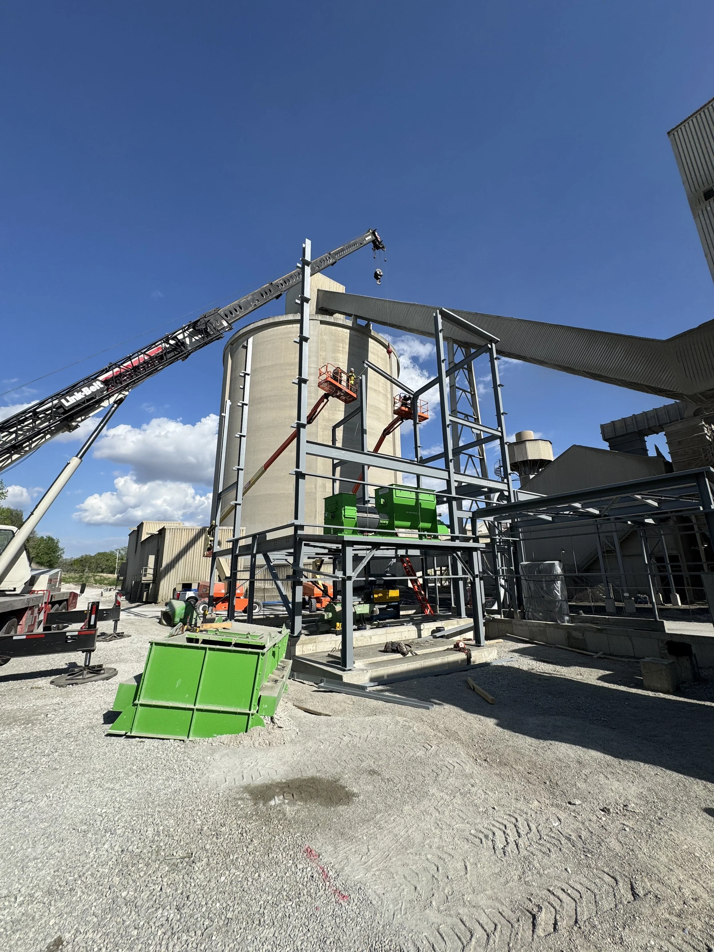 Industrial construction site with steel framework, a crane, green equipment, and a large silo under a blue sky.