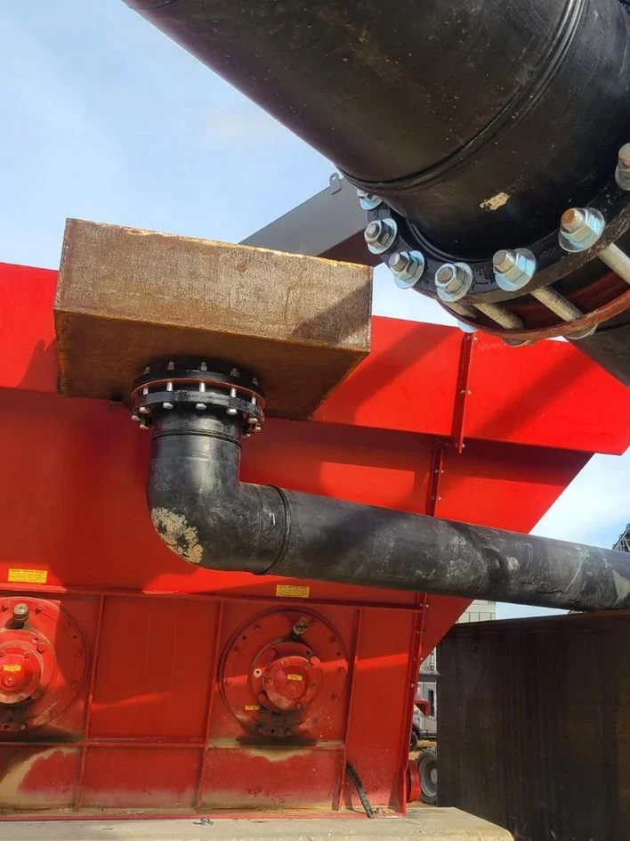 Close-up of industrial piping and equipment, featuring large black pipes, bolted connections, and a red metal structure supporting the pipes at an outdoor industrial site.