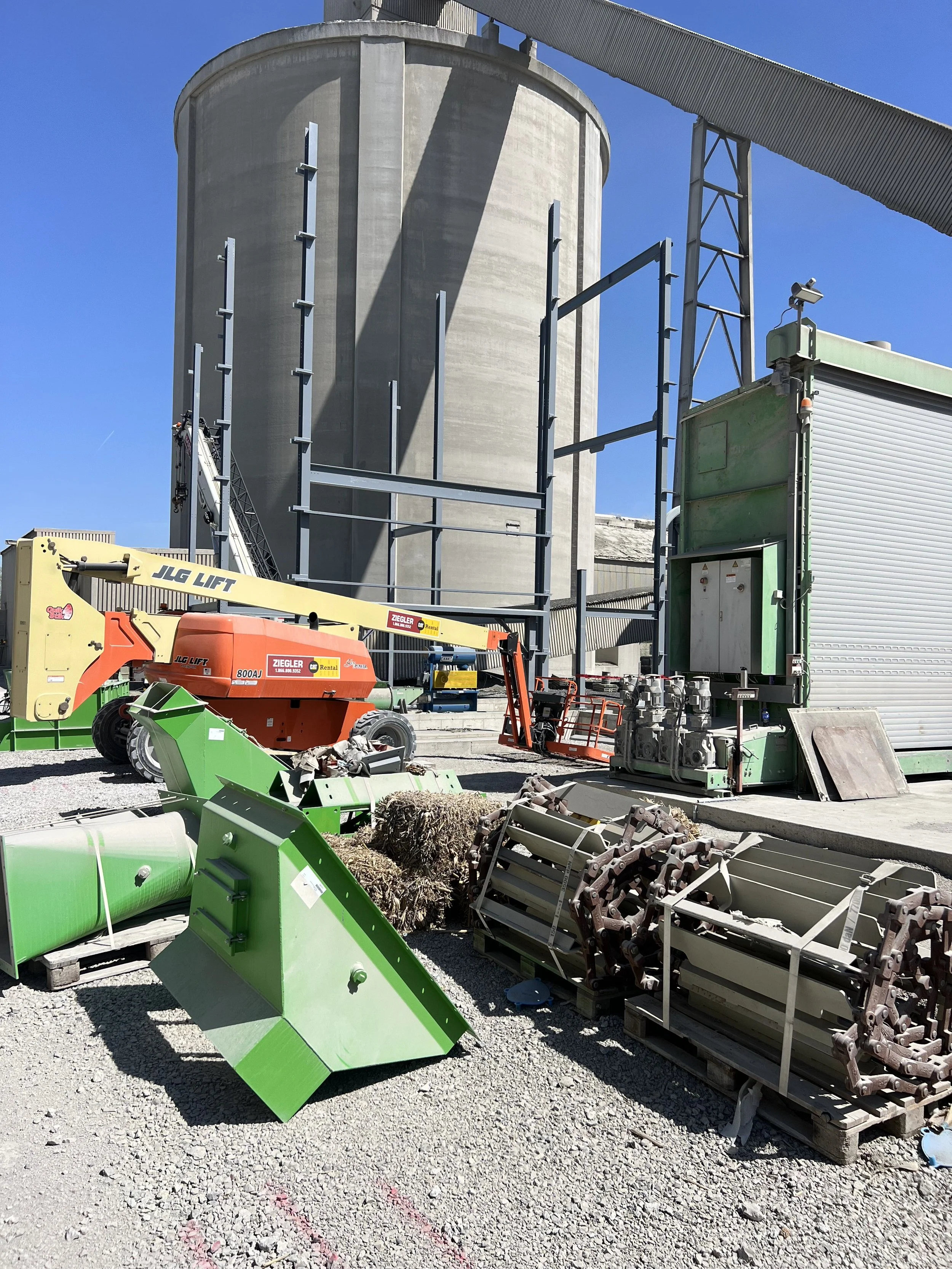 Construction site with various equipment and materials, including green and orange machinery, metal frames, and a large concrete silo against a blue sky.