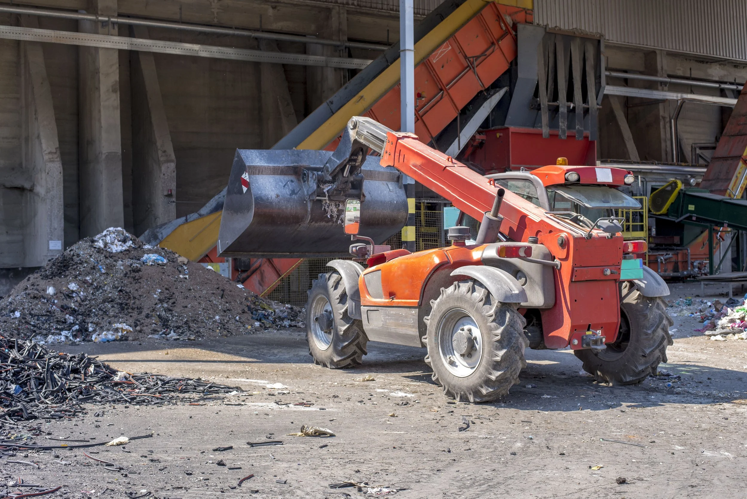 A construction or waste disposal site with a large orange front loader, a pile of mixed debris, and machinery in the background under a concrete structure.