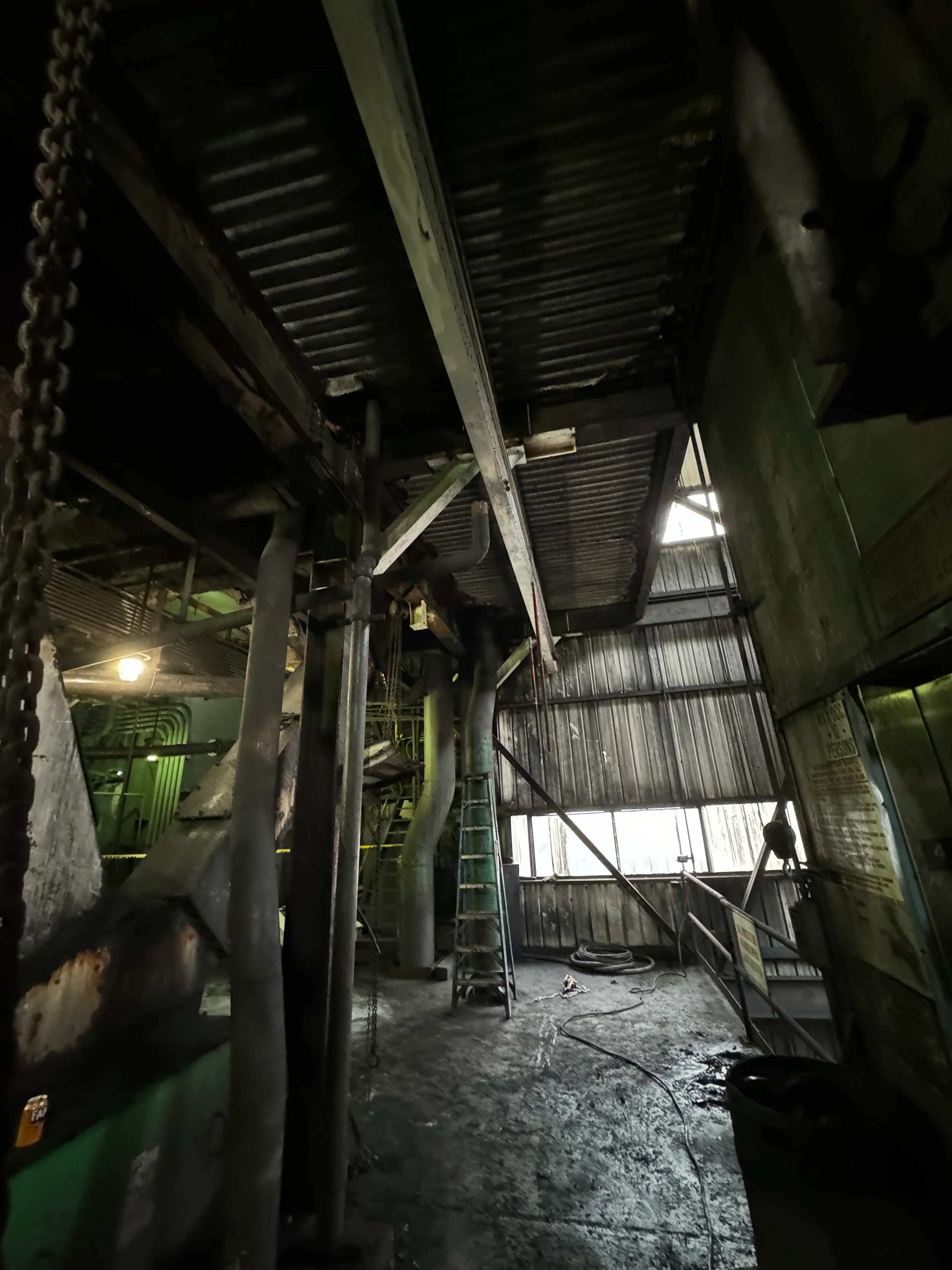 Industrial workshop with metal scaffolding, pipes, a ladder, and a wet concrete floor. Dim lighting, with some safety and warning signs on the wall.