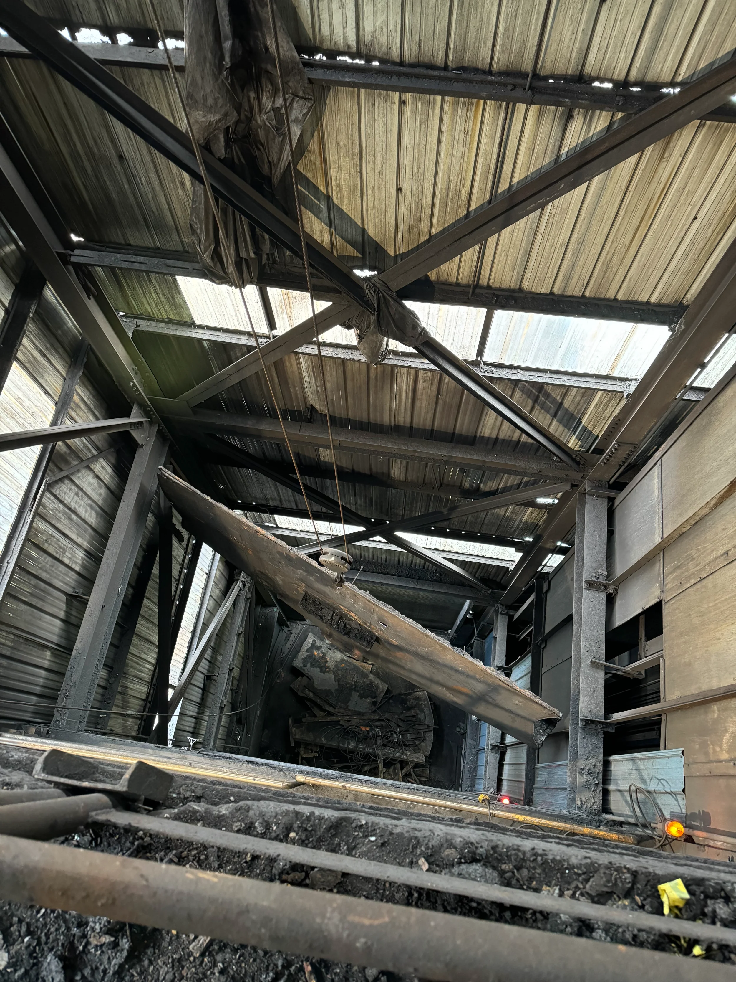 The image shows a building's structure after a fire, with charred and damaged metal beams, burnt roofing materials, and debris on the ground. The roof has holes with sunlight shining through.