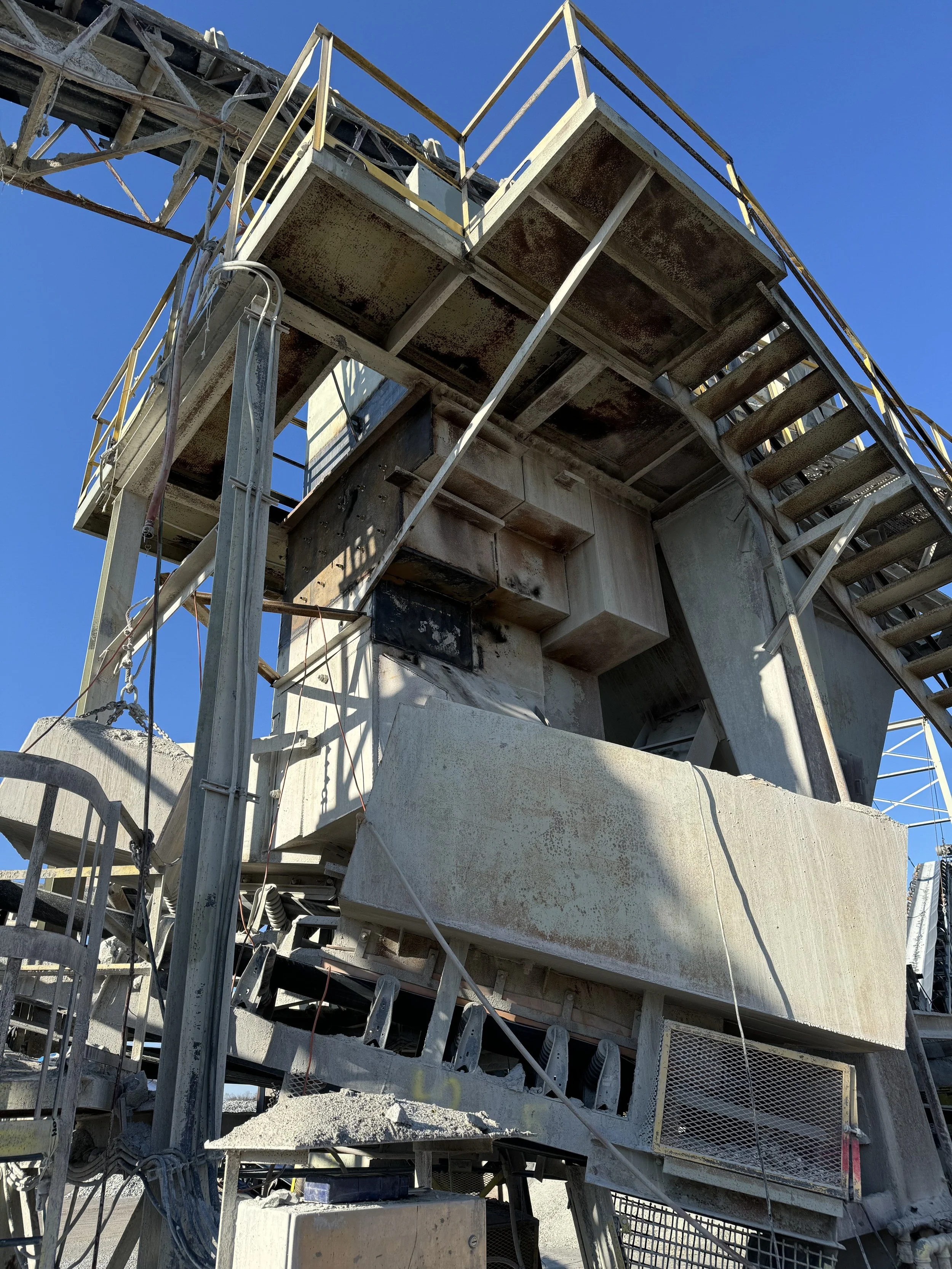 Close-up view of a large industrial concrete and metal structure, possibly part of a construction site, with rusted steel and concrete components, against a clear blue sky.