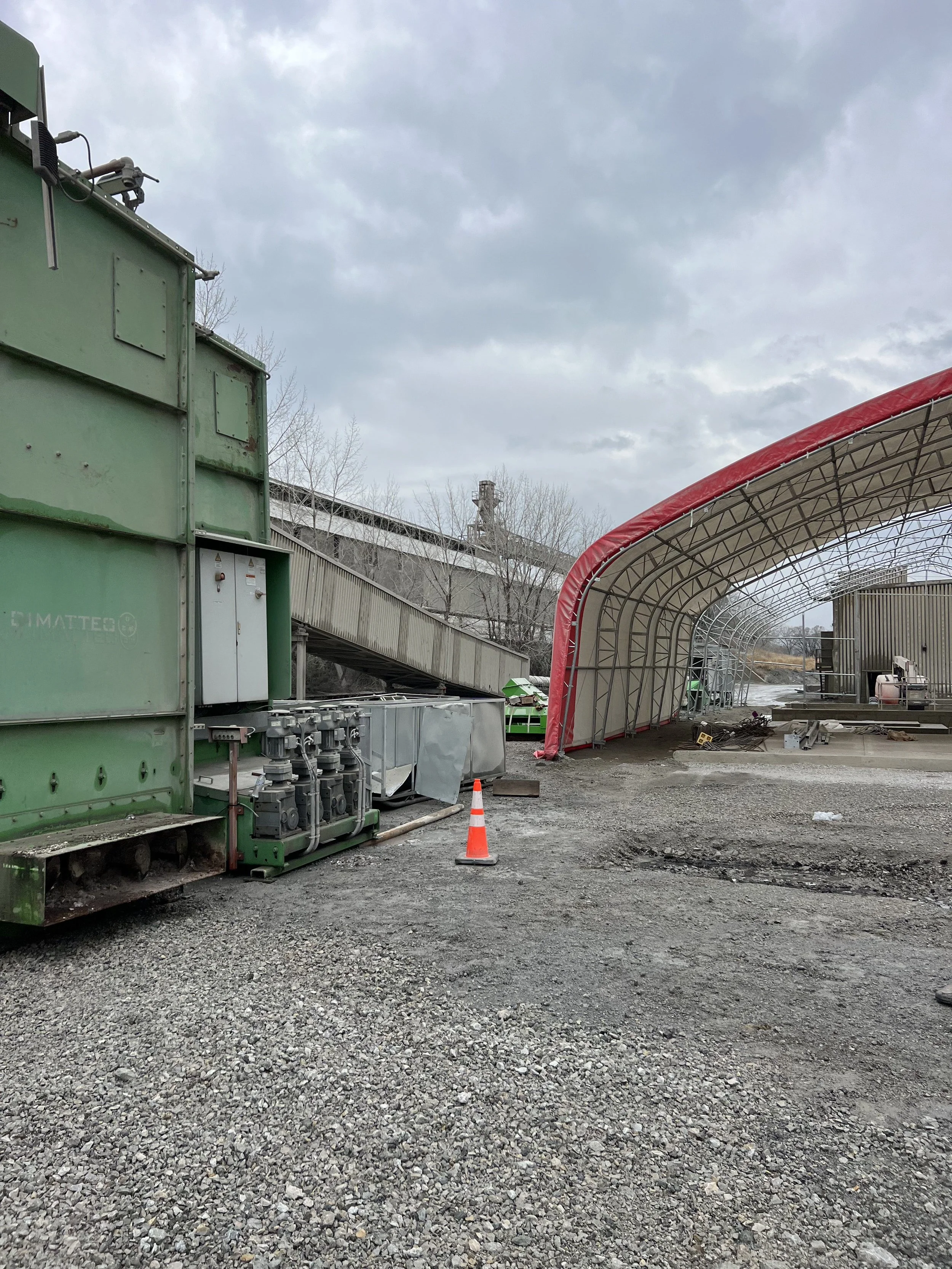 Construction site with gravel ground, green equipment on the left, a red and gray tunnel-like structure on the right, and a cloudy sky overhead.
