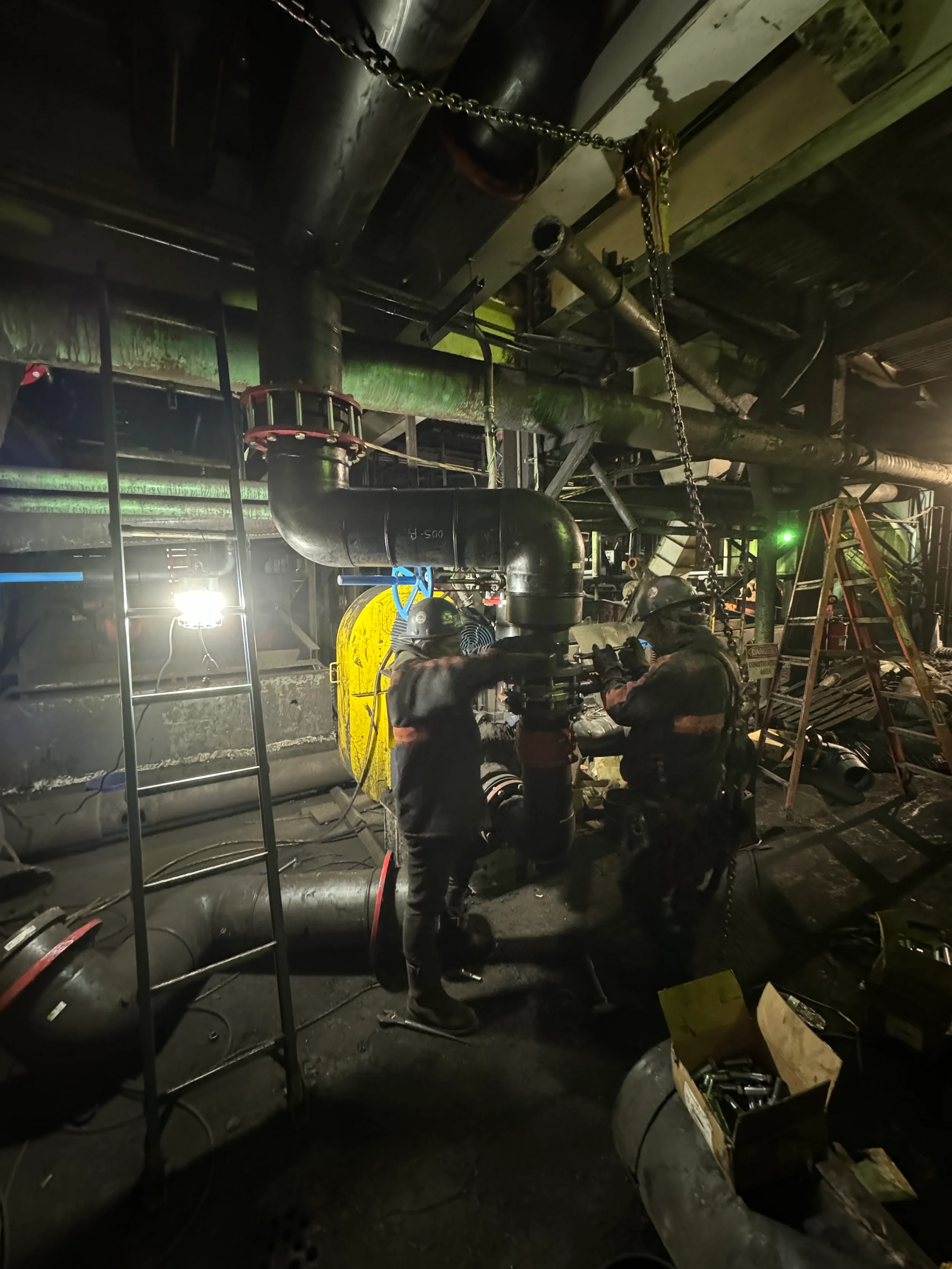 Workers are cutting and assembling pipes in a dimly lit industrial setting, with tools, ladders, and equipment around.