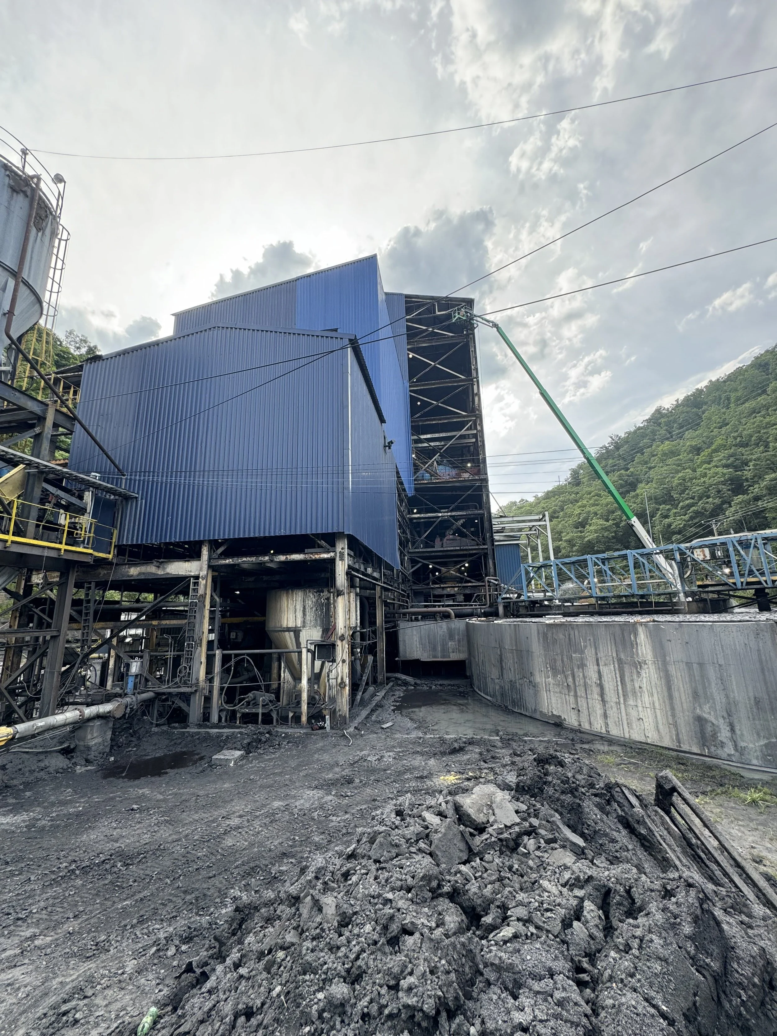 Construction site with blue buildings, a crane, and scattered rocks and debris on the ground, with green hills and a partly cloudy sky in the background.