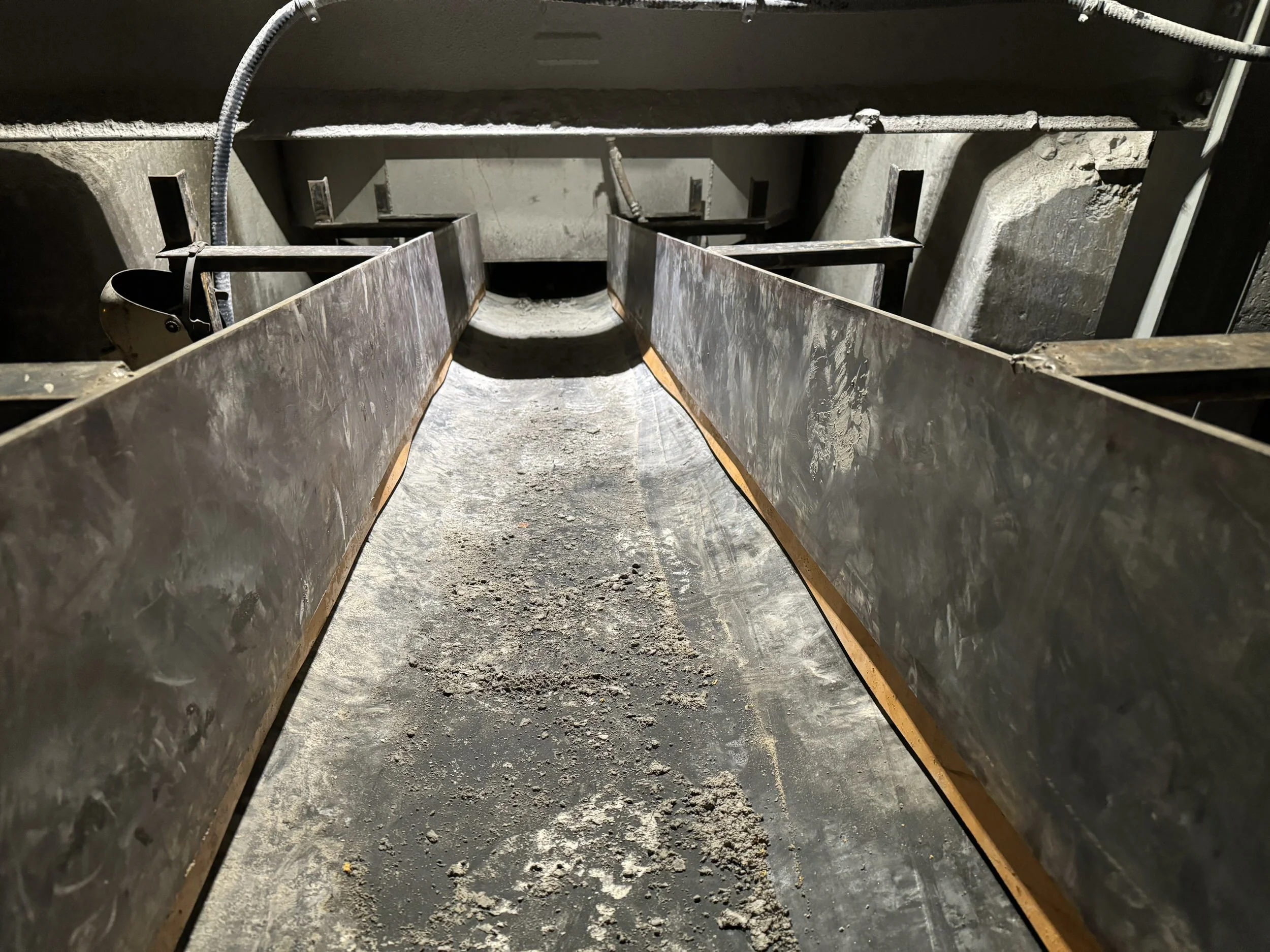 Looking into a tunnel or sewer with metal walls and a dirt floor, showing a section of a large underground space, possibly for cables or pipes, with some wires or hoses visible on the walls.