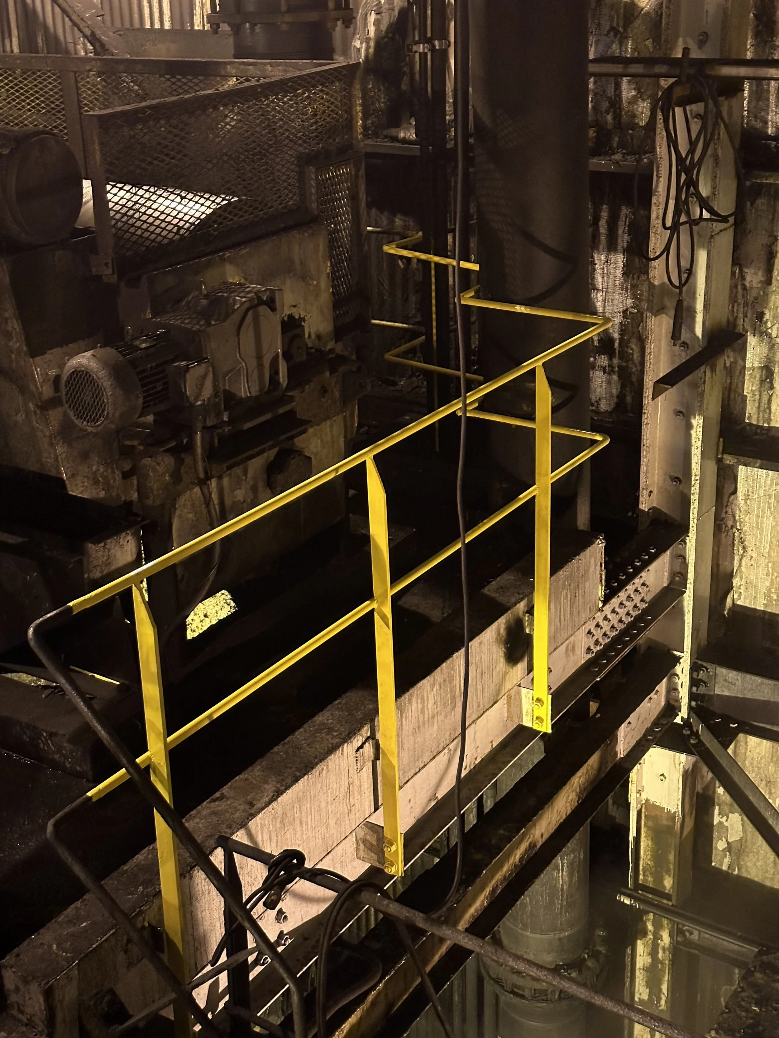 View of an industrial underground tunnel or mining shaft with machinery, yellow safety rails, pipes, and walls covered in soot and grime, illuminated by overhead lighting.