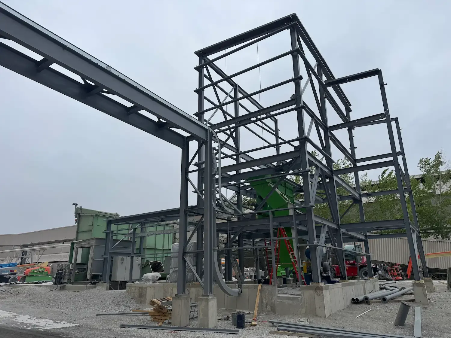 Construction site with steel framework for a building, some equipment and materials on the ground, and a cloudy sky.