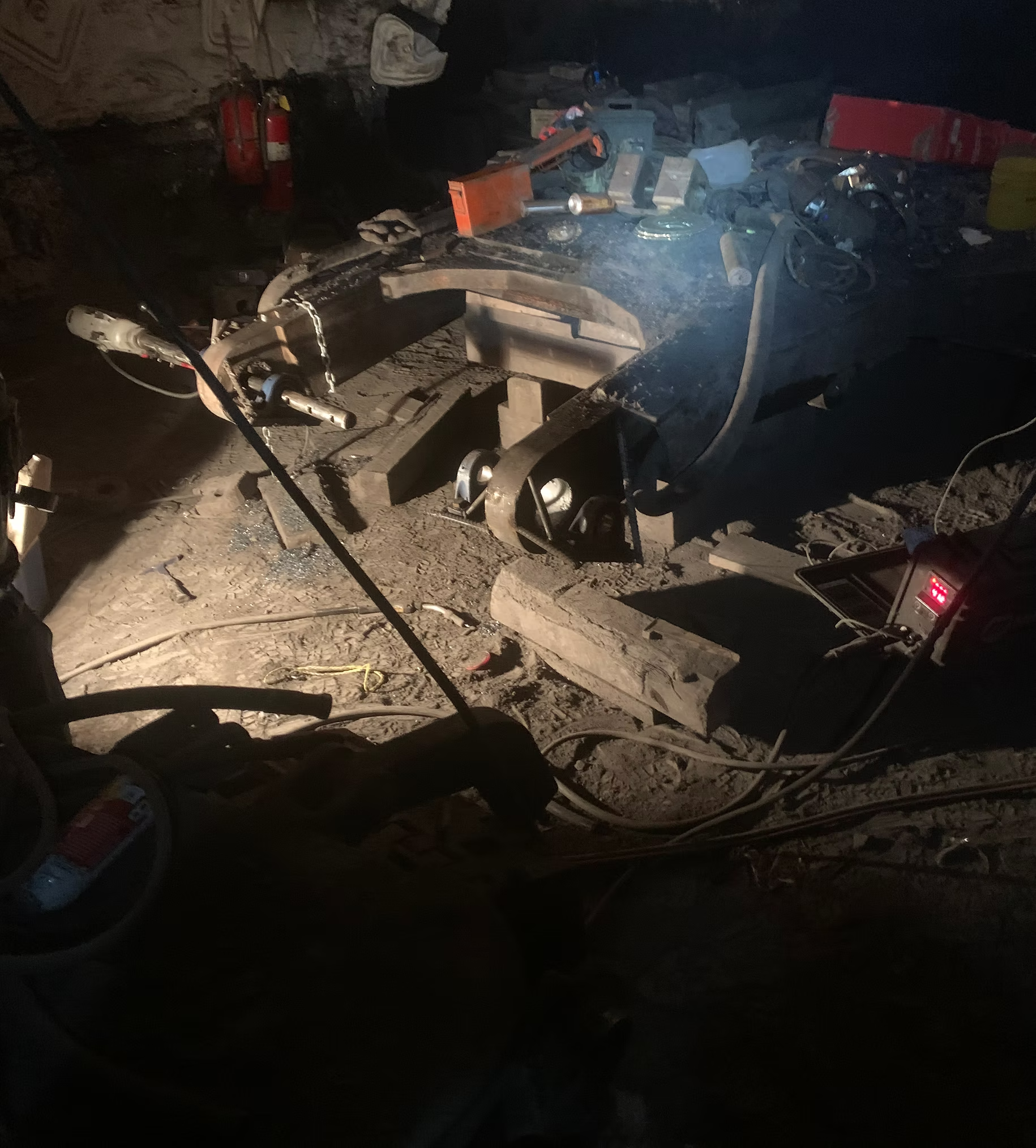 A construction or work site with scattered tools, equipment, and debris. Includes fire extinguishers, a fire alarm, wires, and various construction materials on the ground and surrounding area.