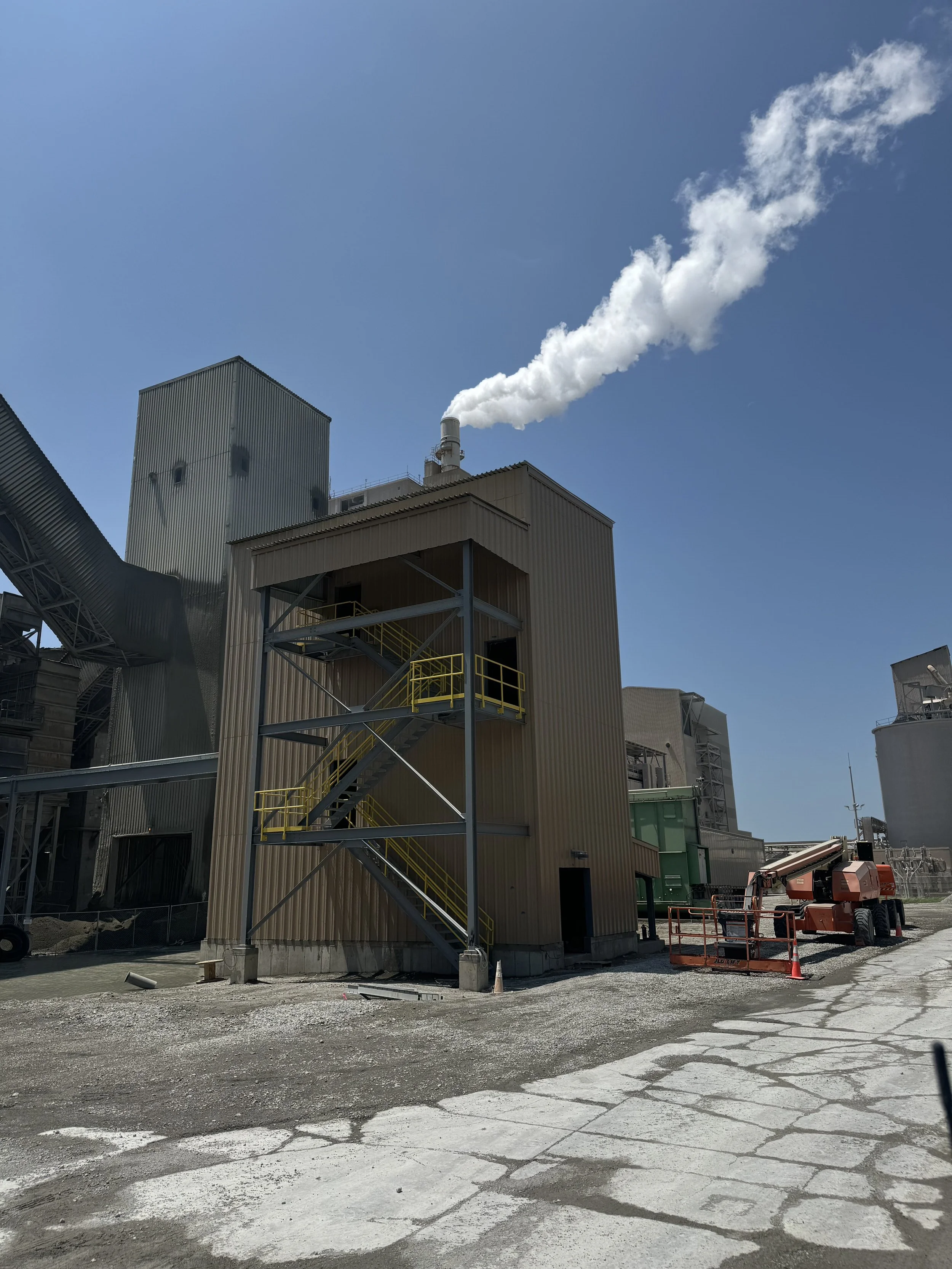 Construction site with industrial building, metal stairs, and a crane on a partly paved ground, emitting white smoke or steam into a clear blue sky.