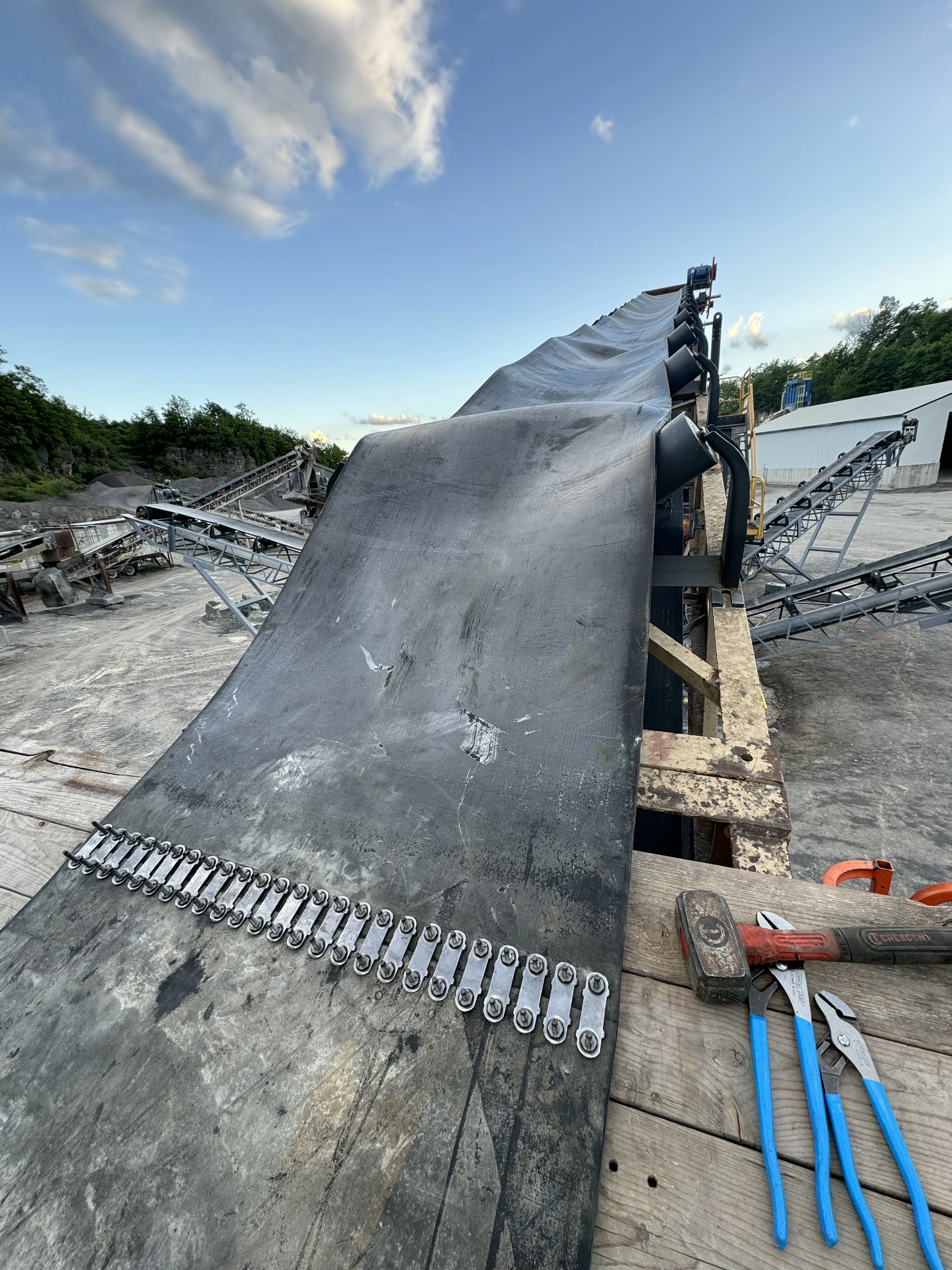 Construction site with conveyor belt, cutting tools, and construction tools on a wooden plank, under a blue sky with clouds.