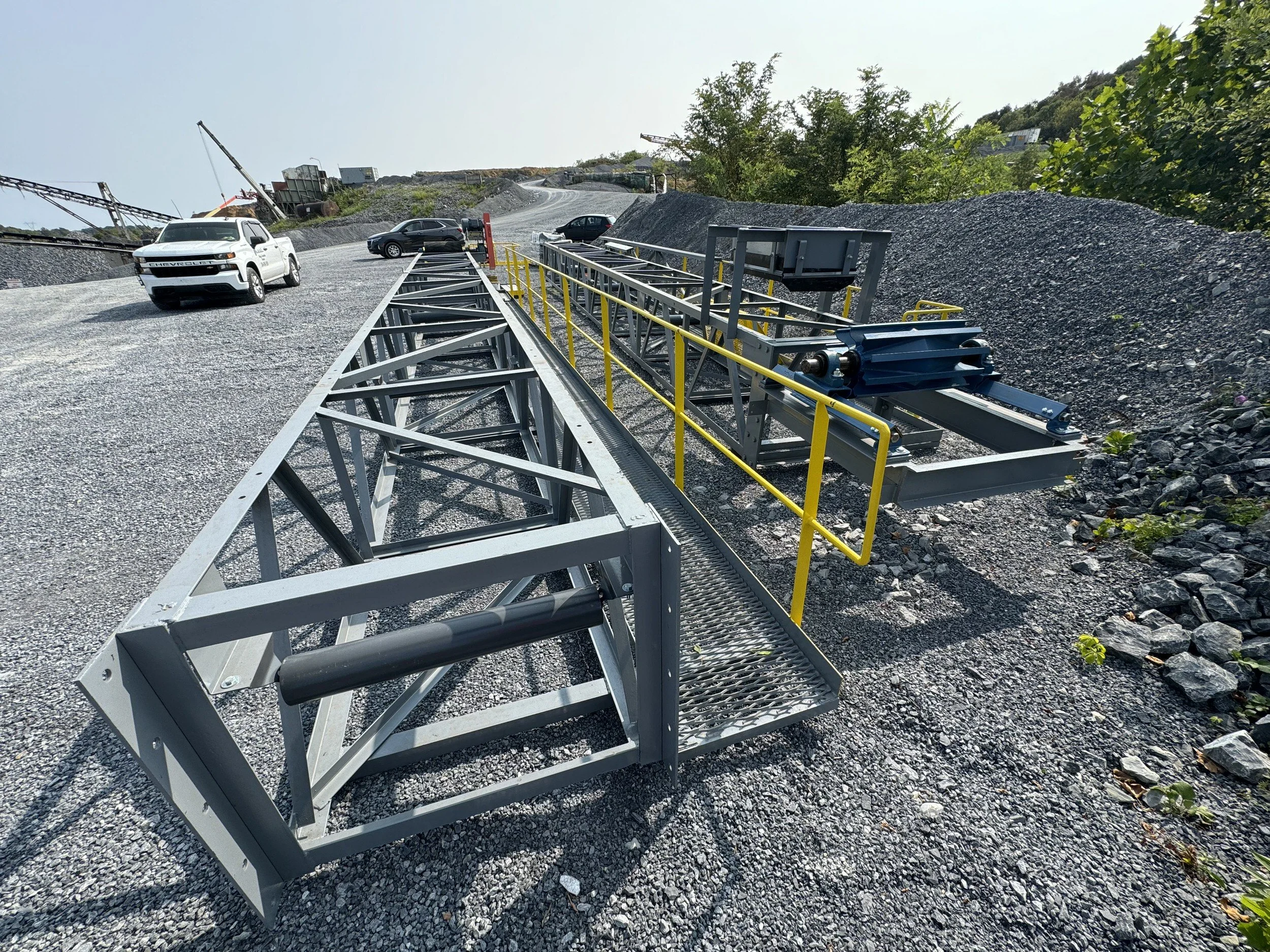 A construction site on a hillside with parked cars, gravel, and a metal roller machine. In the background, there are trees and construction equipment.