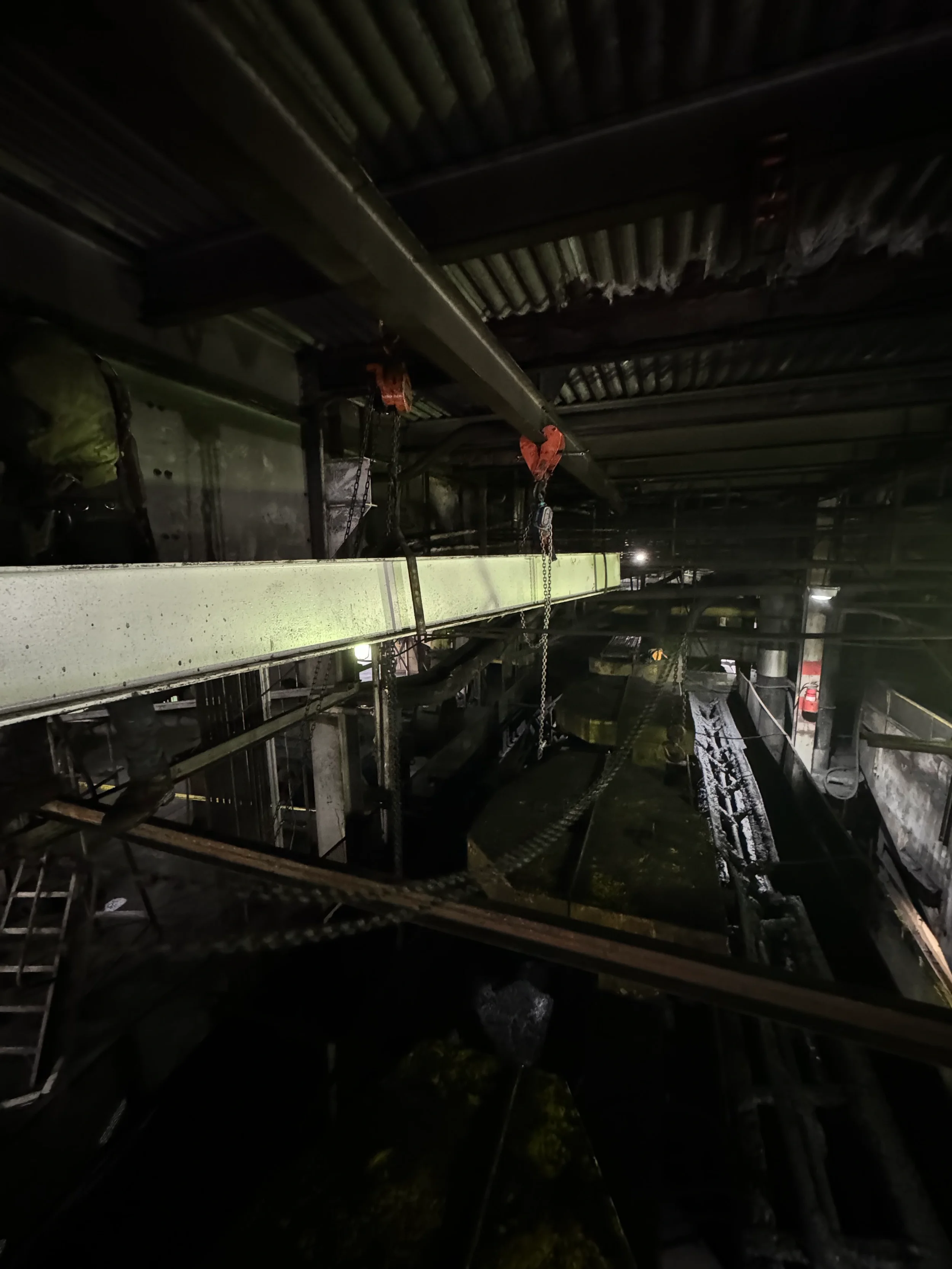 Dark interior of a factory or industrial space with metal beams, chains, and machinery, illuminated by faint lighting.