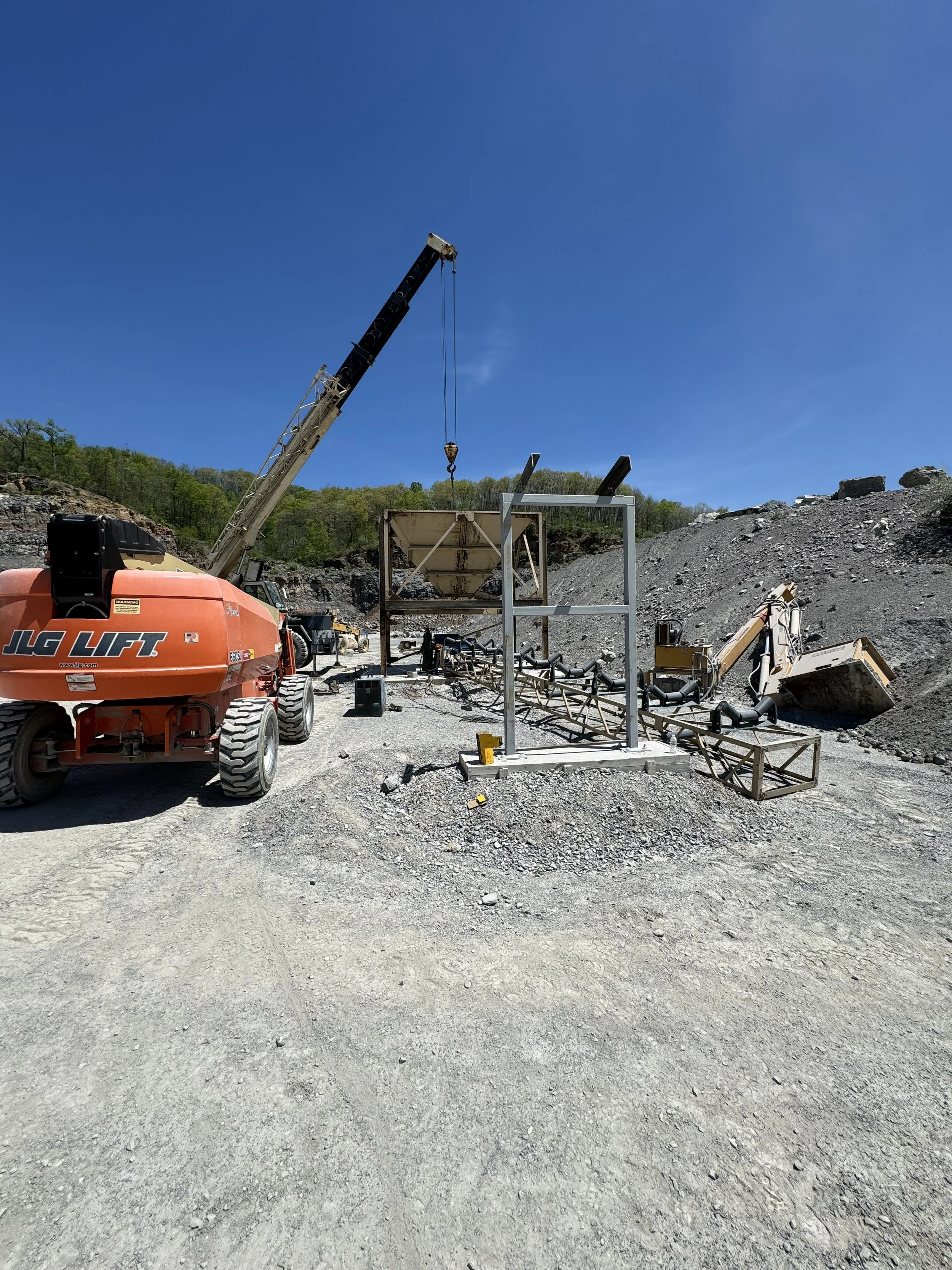 Construction site with a crane lifting a large object, section of a rocky hill, and construction equipment on a dirt ground.