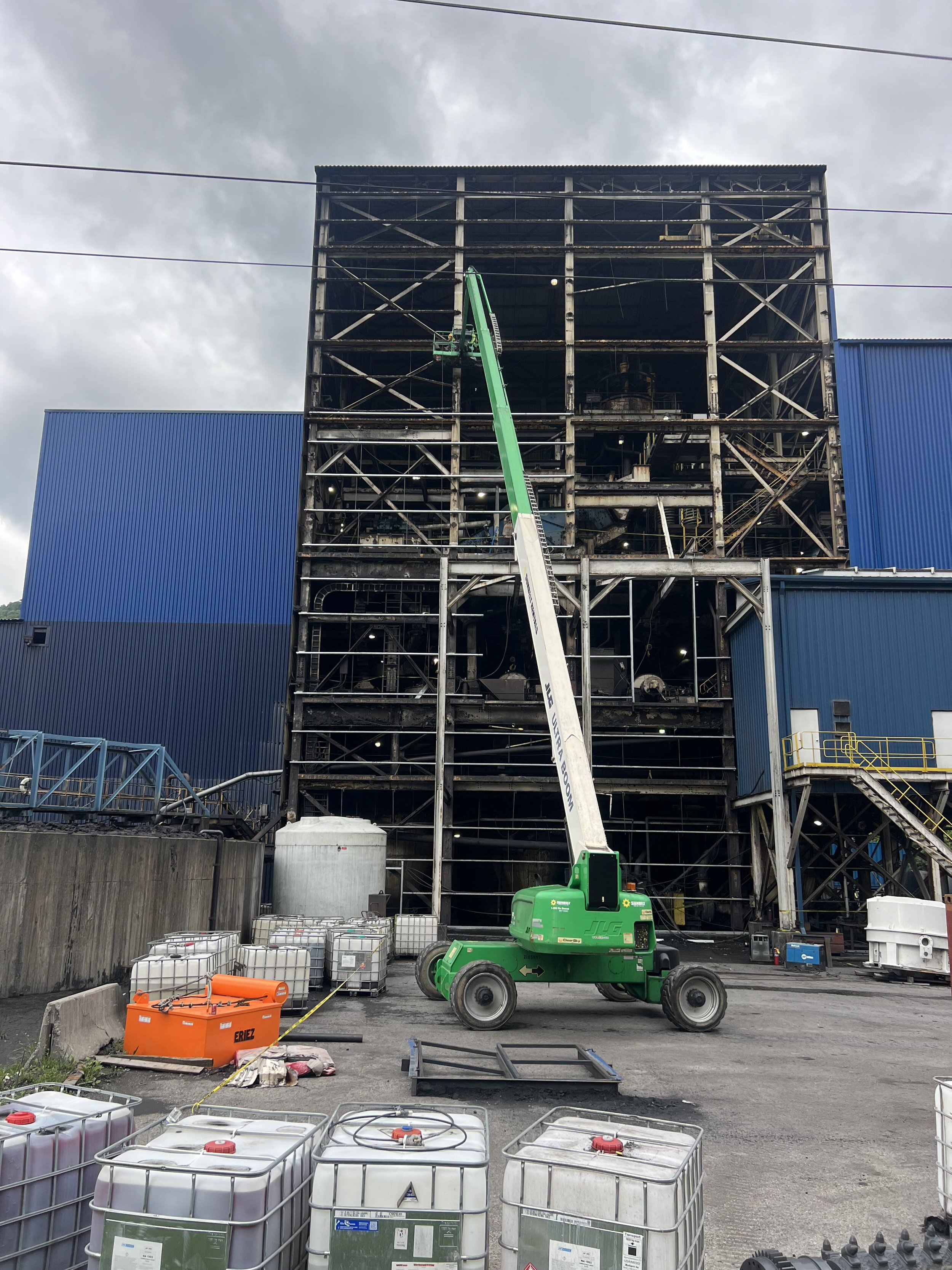 Construction site with a steel framework building under renovation, a green lift elevating workers, blue metal siding on parts of the structure, and various containers and equipment on the ground.