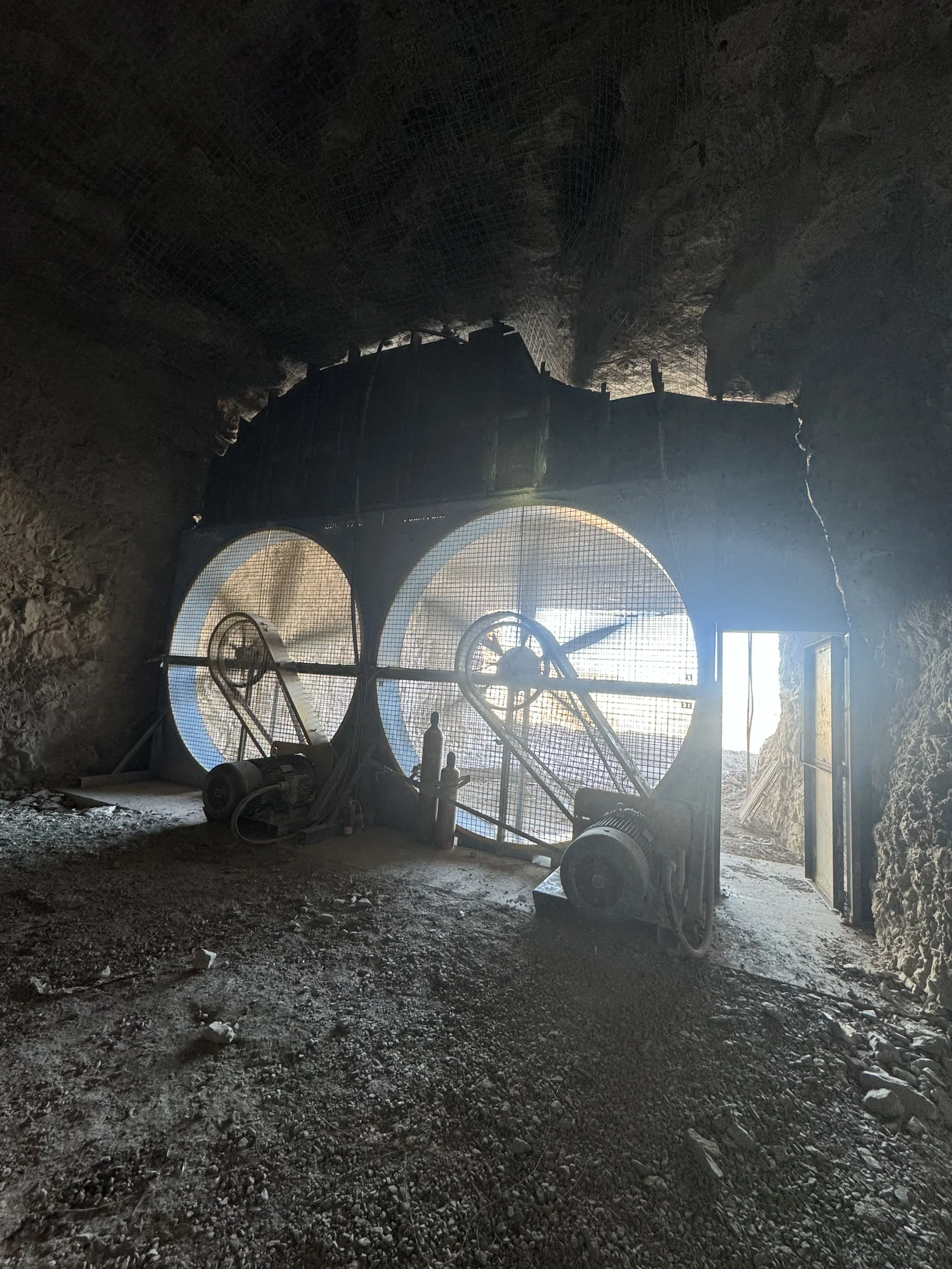 Underground mining tunnel with large ventilation fan, metal safety barriers, and rocky walls.