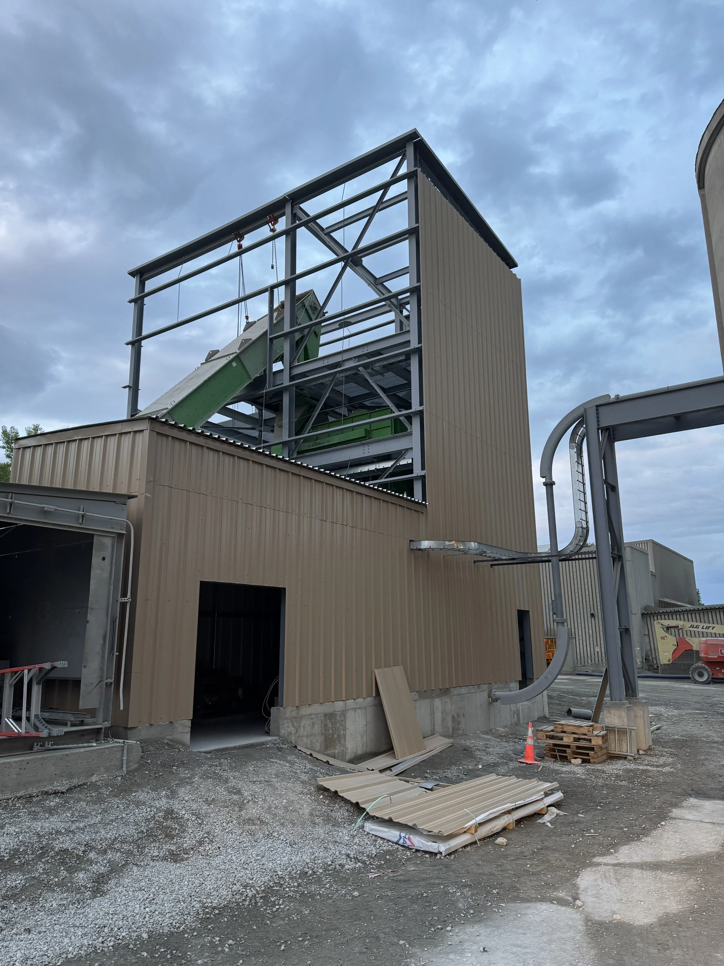 Construction site with partially built commercial building, metal framework, and roofing materials. Gravel and dirt ground with some construction equipment and debris.