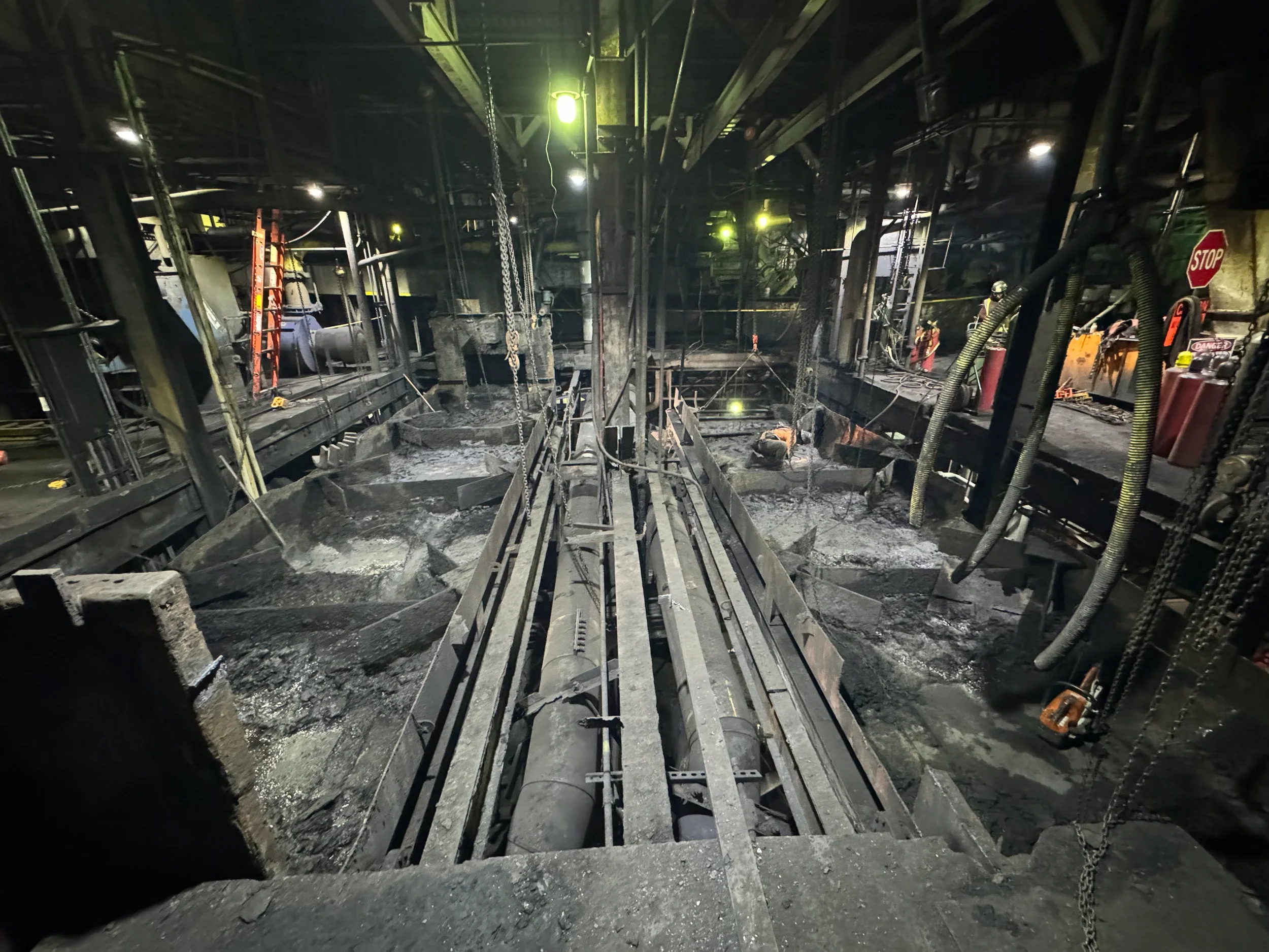 Interior of a construction site with steel framework, chains, and construction workers in safety gear and helmets amid debris and industrial equipment.
