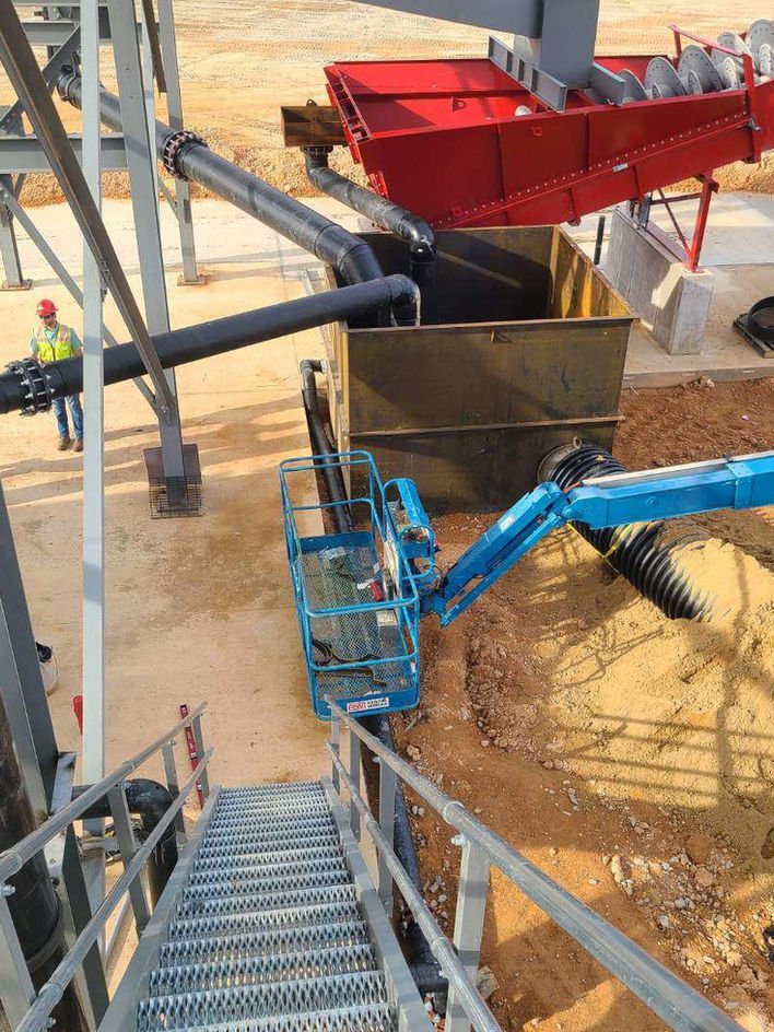 Construction site with large pipes and machinery including a blue lift, red conveyor, and dirt in-ground area with some workers nearby.