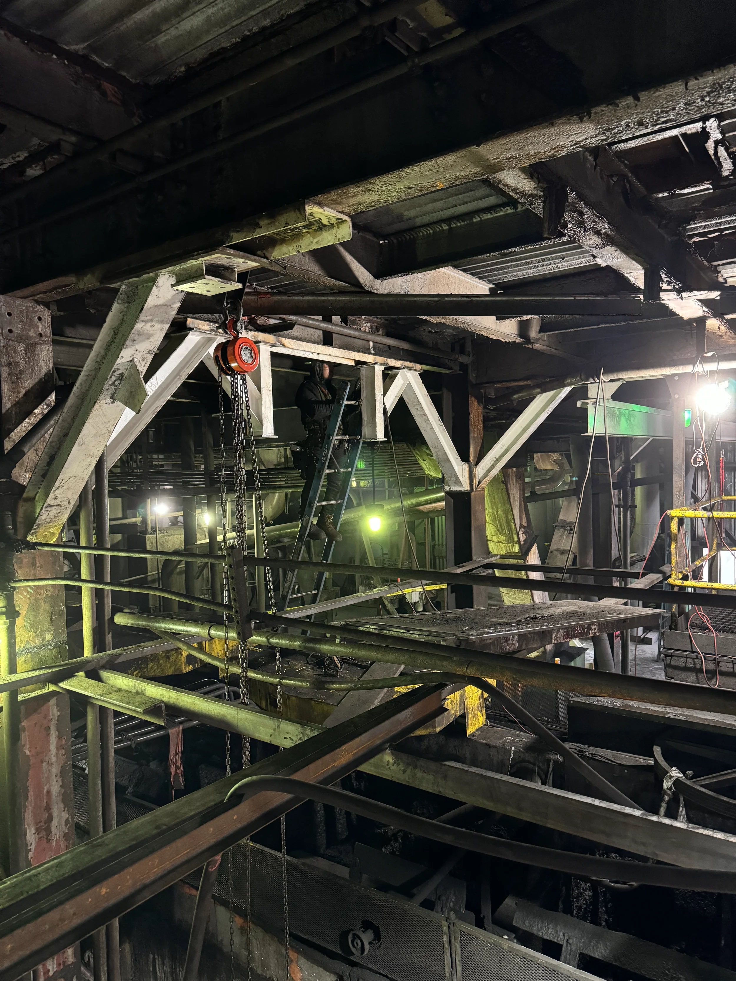Underneath a large industrial or construction site structure, showing metal scaffolding, pipes, chains, and a worker standing on a ladder working on the infrastructure.