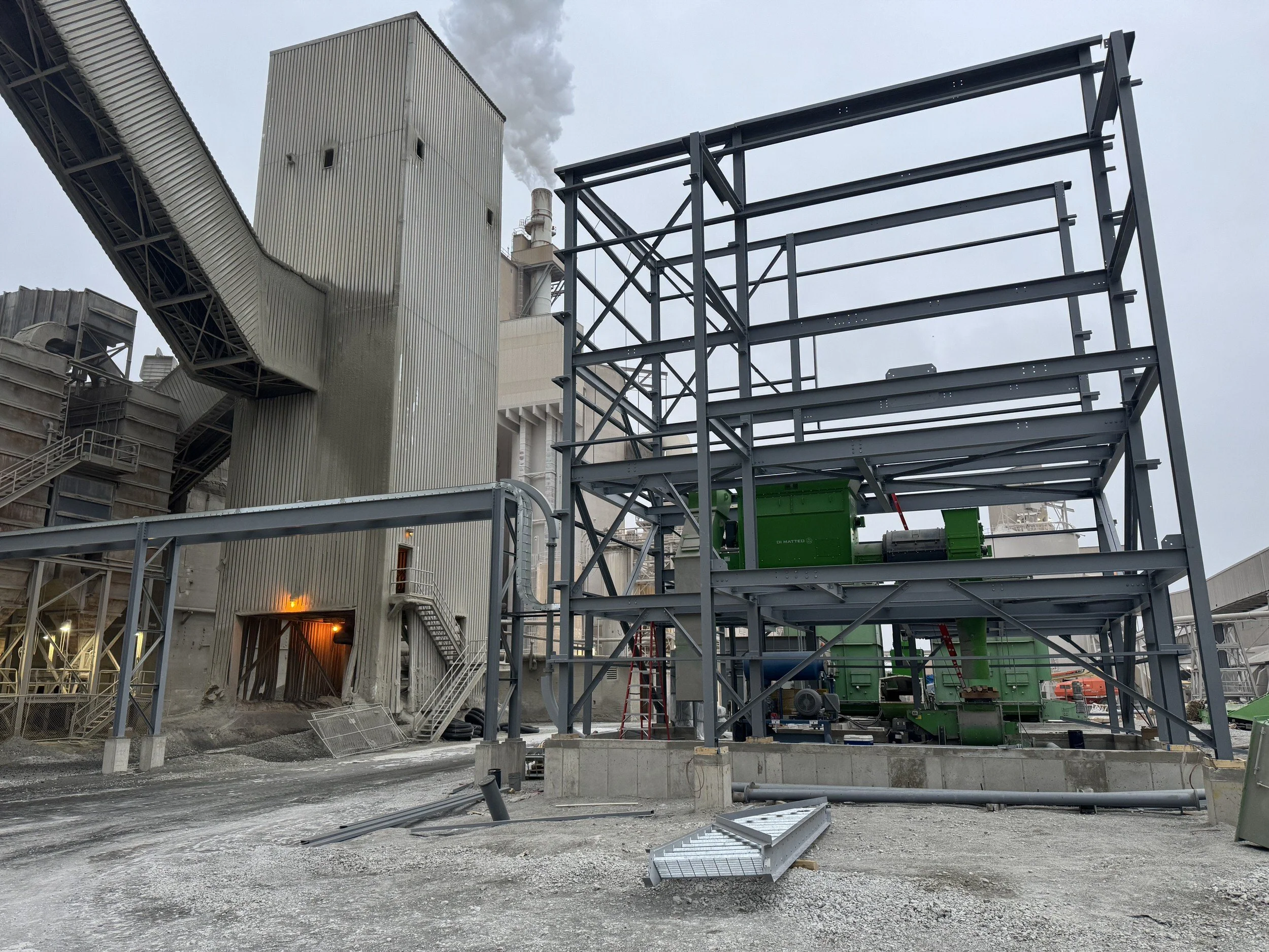 Industrial construction site with steel framework, large piping, and machinery under cloudy sky.