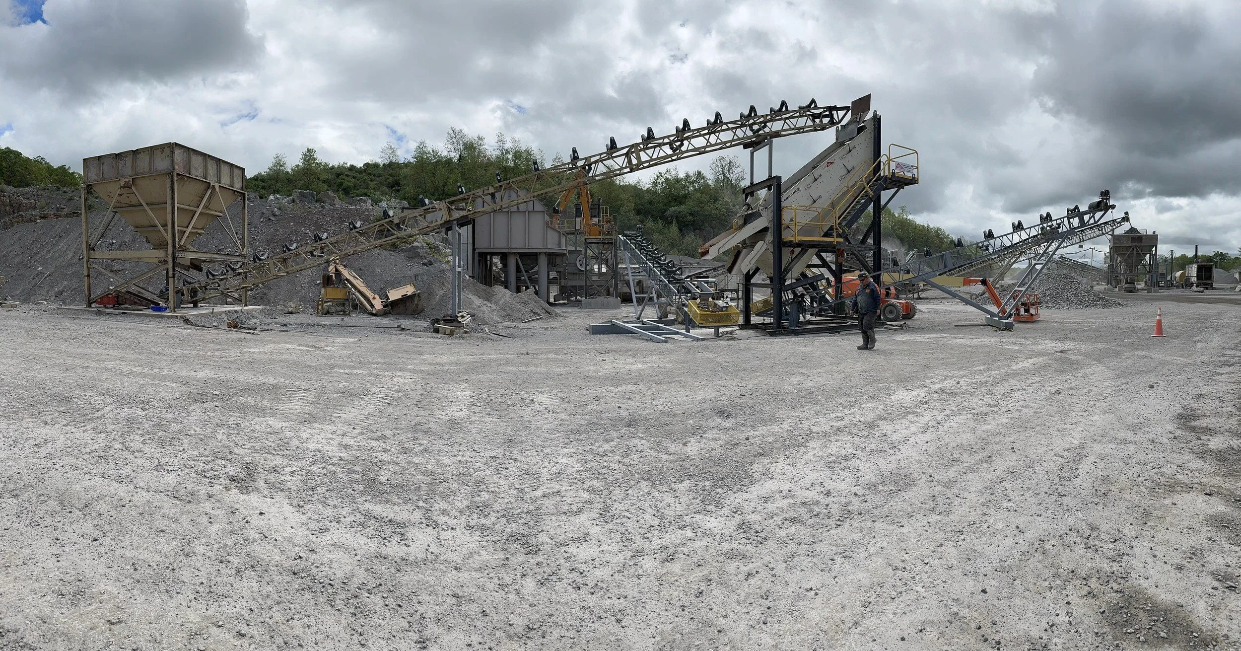 A gravel quarry or construction site with multiple conveyor belts, machinery, and workers, under cloudy skies.