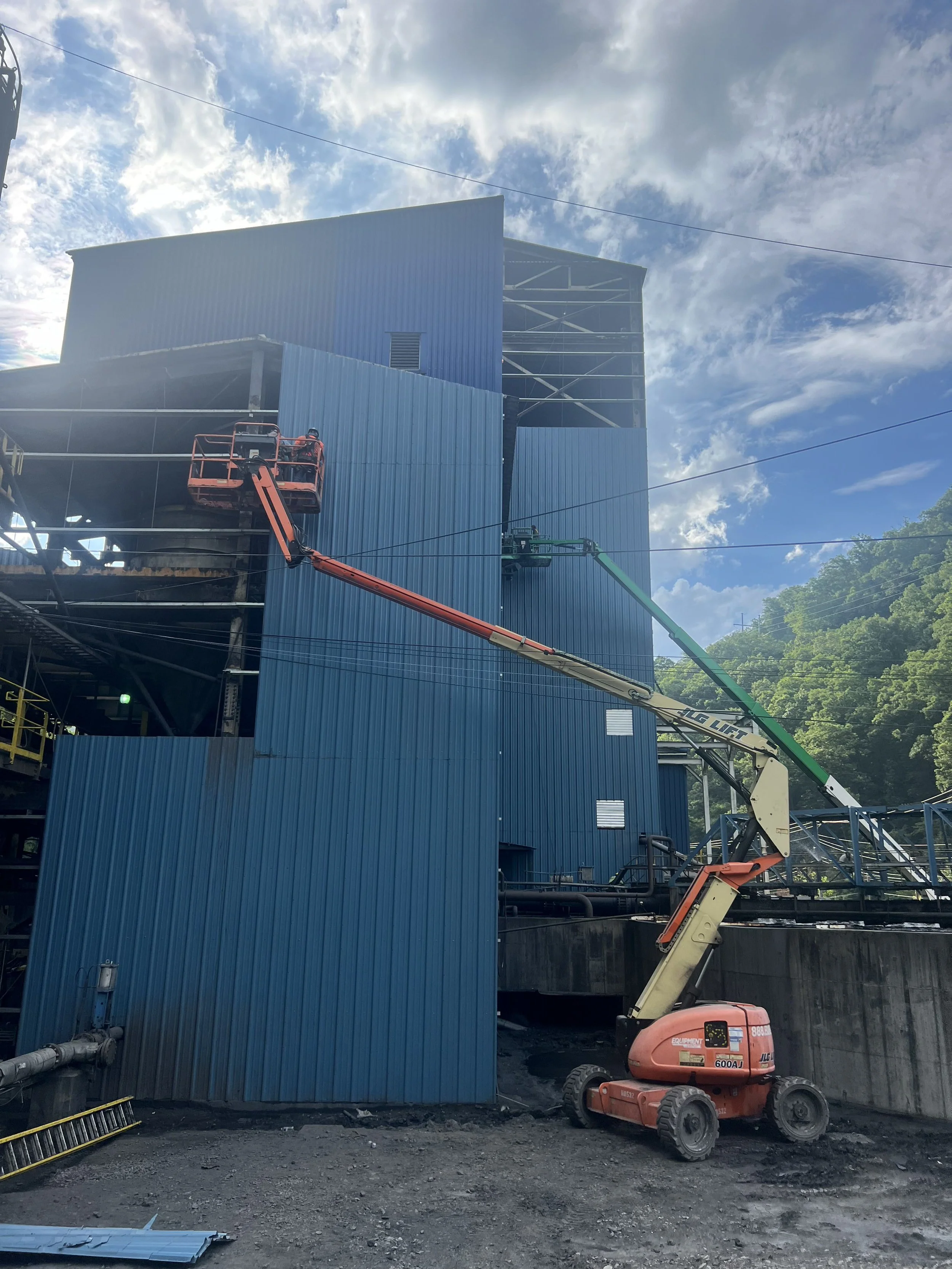 Construction site with a blue metal building, worker on an elevated lift, and a cherry picker with a blue sky and clouds in the background.