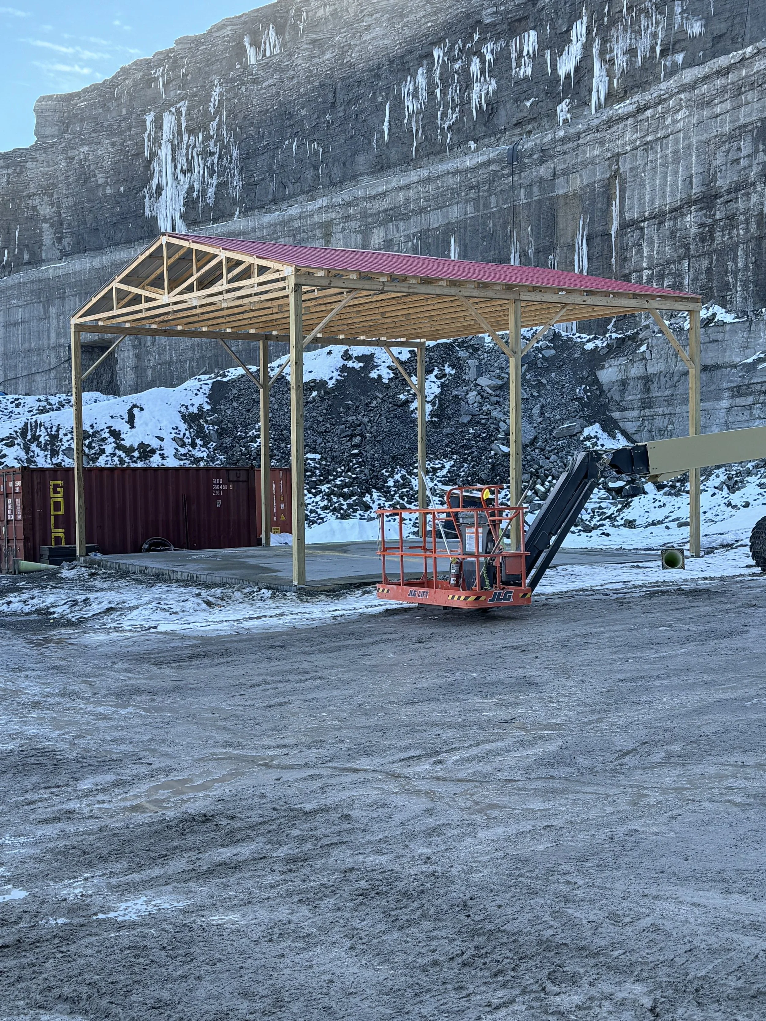 A construction site with a wooden frame structure of a building and a red roof, set against a rocky mountain backdrop with snow and icicles, and a gravel or dirt ground with snow patches.