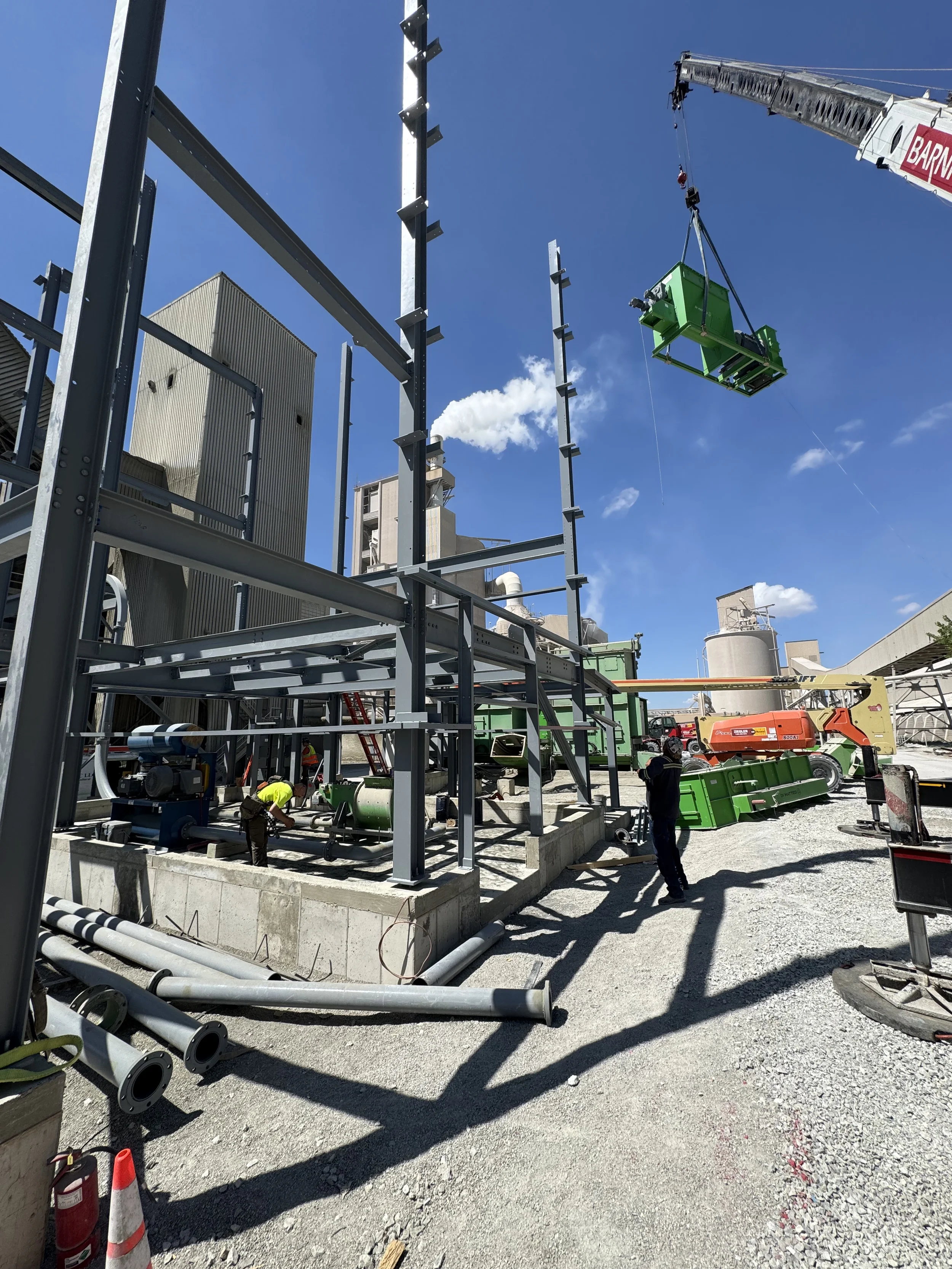 Construction site with steel framework, workers, and a crane lifting a green structure against a blue sky.