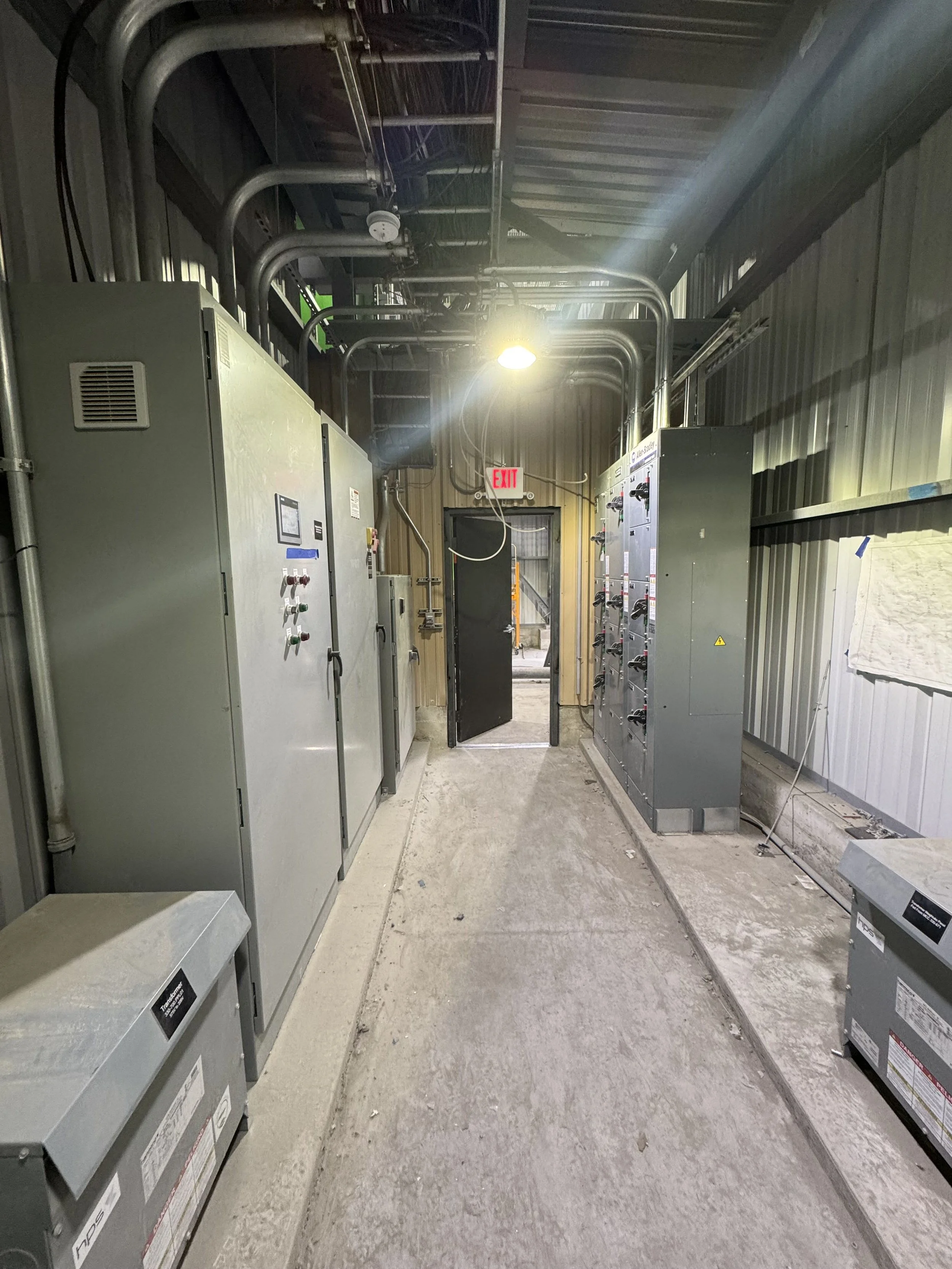 Industrial electrical control panels and piping inside a warehouse or utility room with a door and an exit sign.