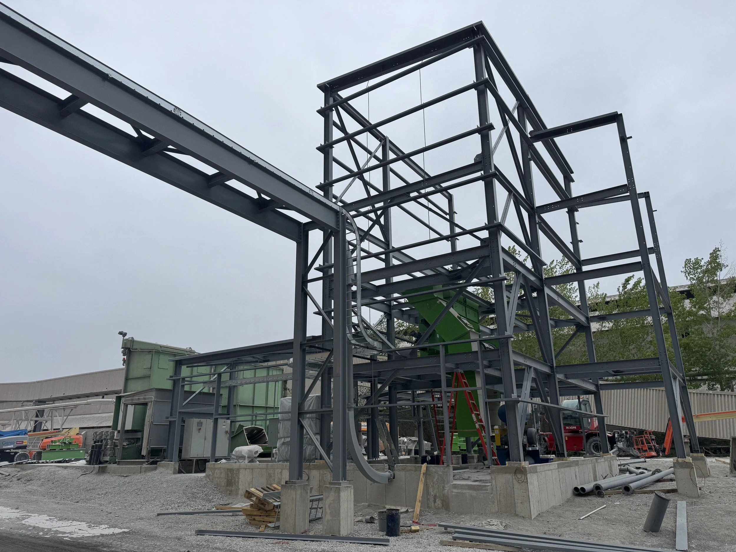 Steel structure under construction with several pipes and equipment, set against an overcast sky and a building in the background.