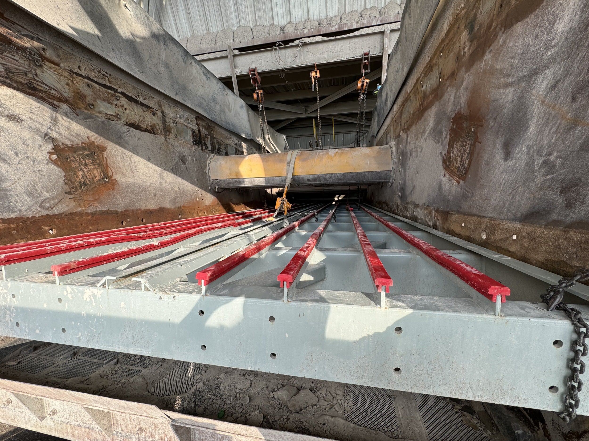 Construction site showing a large trench with concrete walls, red pipes, and metal braces. Equipment and chains are visible at the top and bottom of the trench.