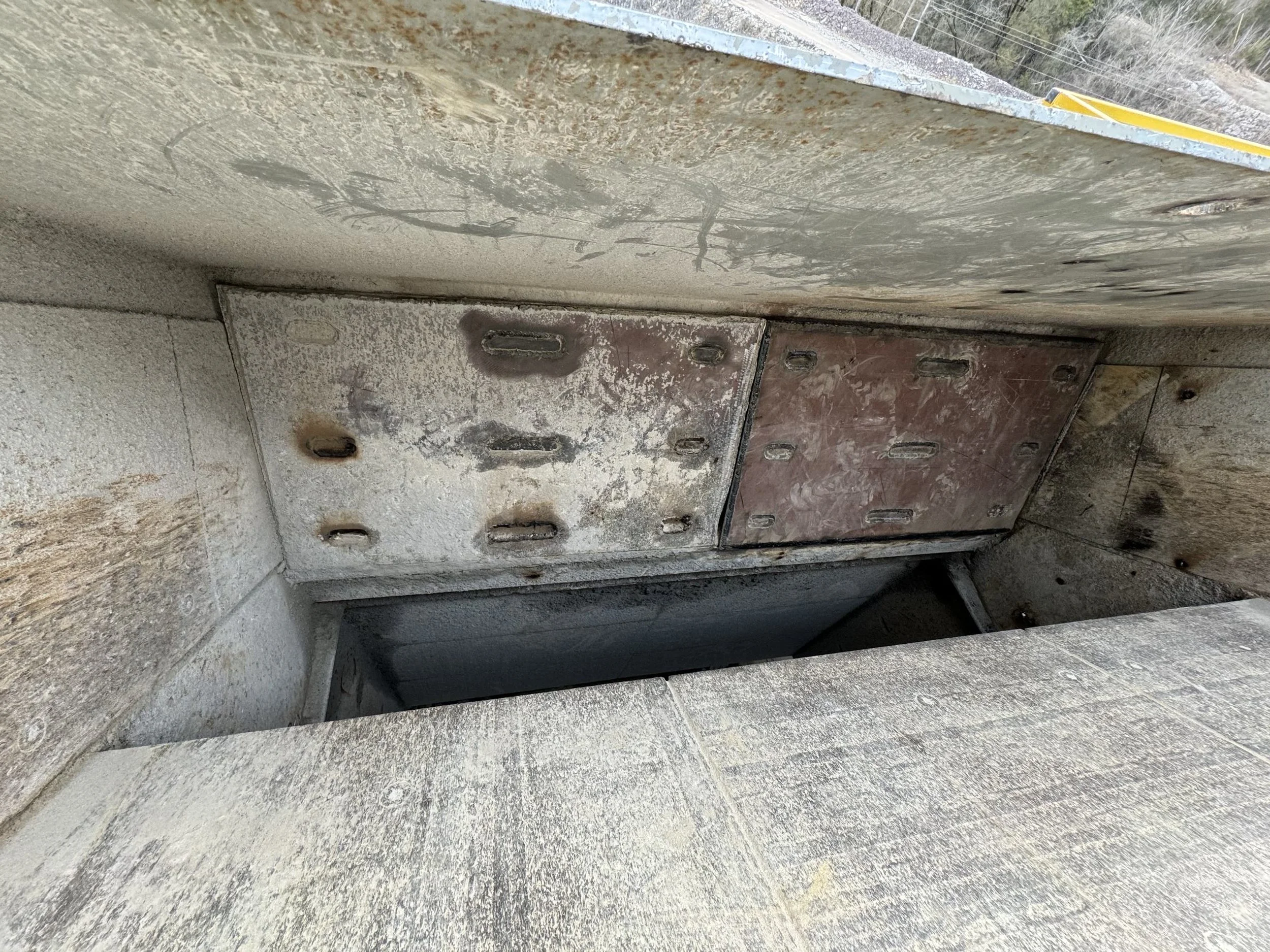 Close-up of a metallic electrical panel or box embedded in a concrete structure, with multiple small openings or holes, some of which are covered with screws or caps, and a dark interior visible through an open slot or door.