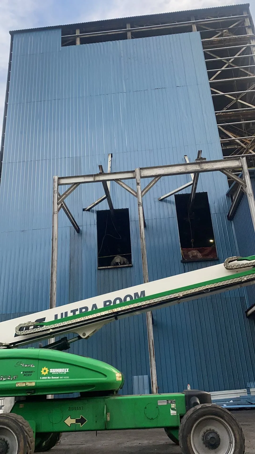 A construction site in front of a large blue building with two openings under construction and metal scaffolding. A green Genie lift with the label 'Sunbelt Rentals' is in the foreground.
