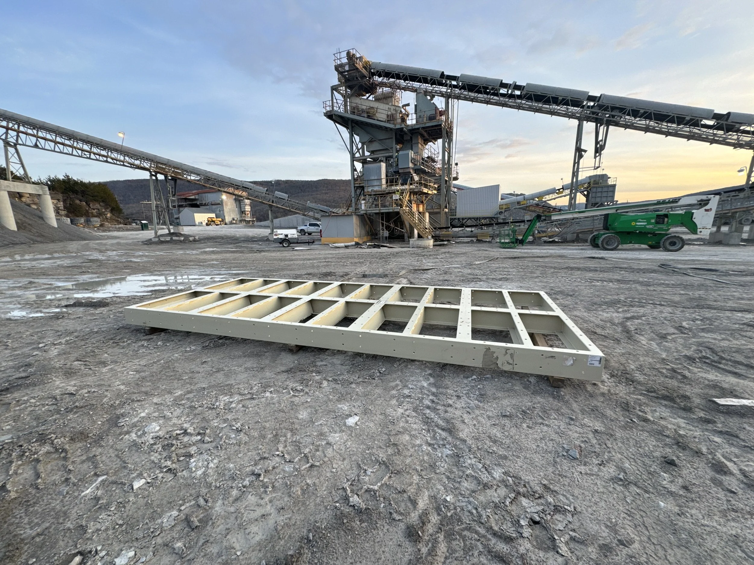 A large industrial mining or construction site with conveyor belts, equipment, and vehicles, set against a backdrop of mountains and a partly cloudy sky.