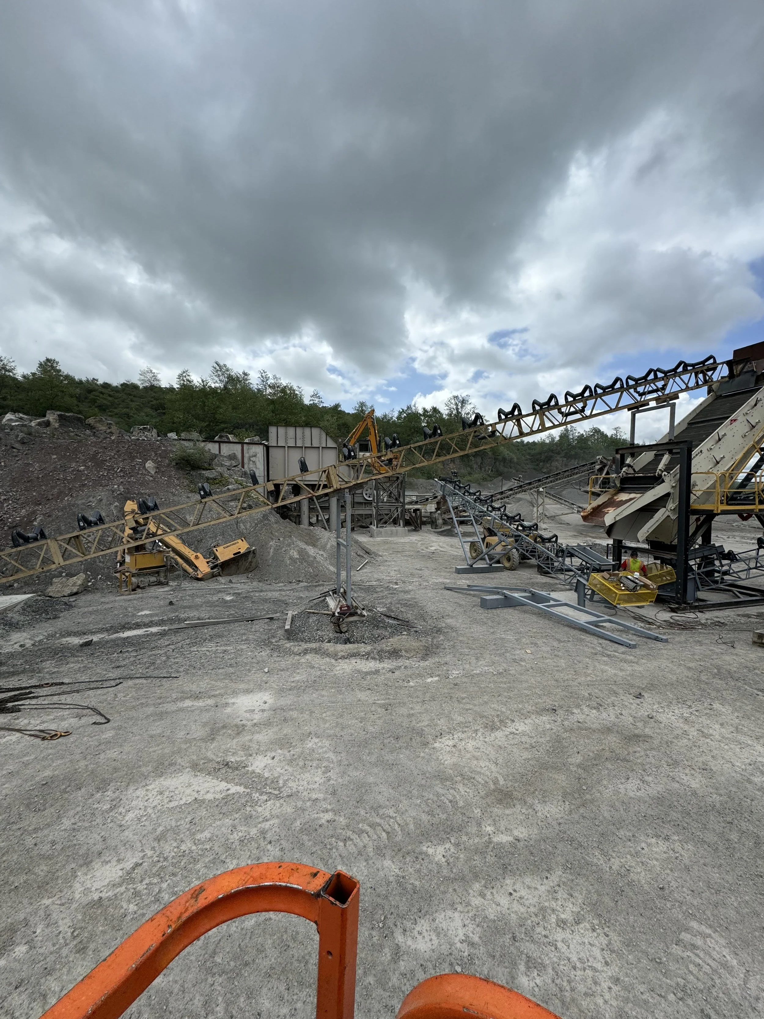 View of a construction site with machinery, conveyor belts, and a dirt ground under a cloudy sky.