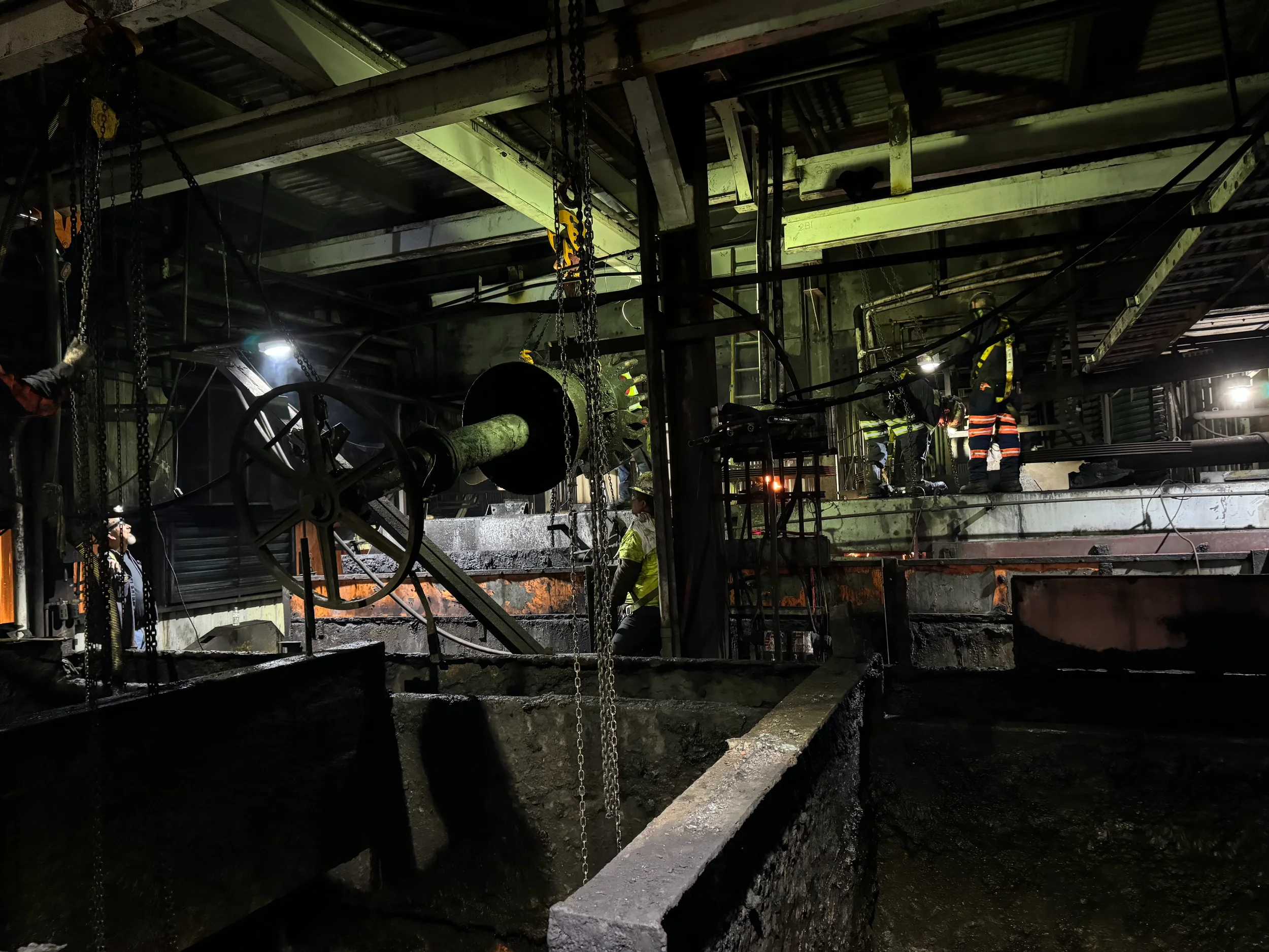 Industrial site with metal beams, chains, and pipes, workers in protective gear, dim lighting, and a smoky atmosphere.