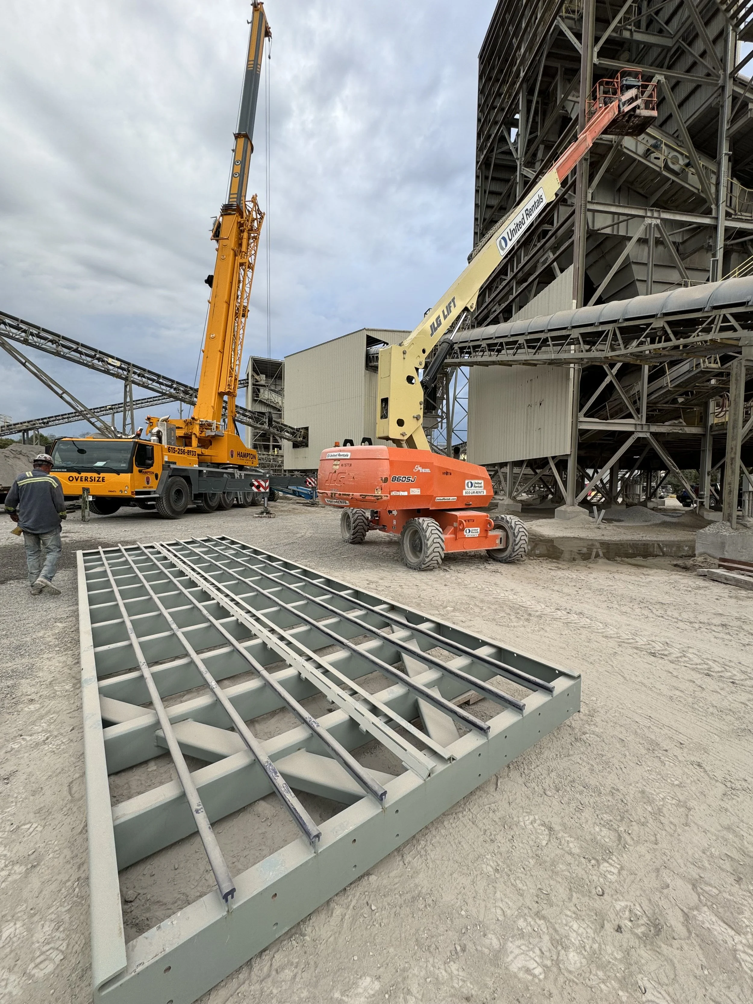 Construction site with cranes, scaffolding, and metal structures, workers, and construction equipment on a cloudy day.